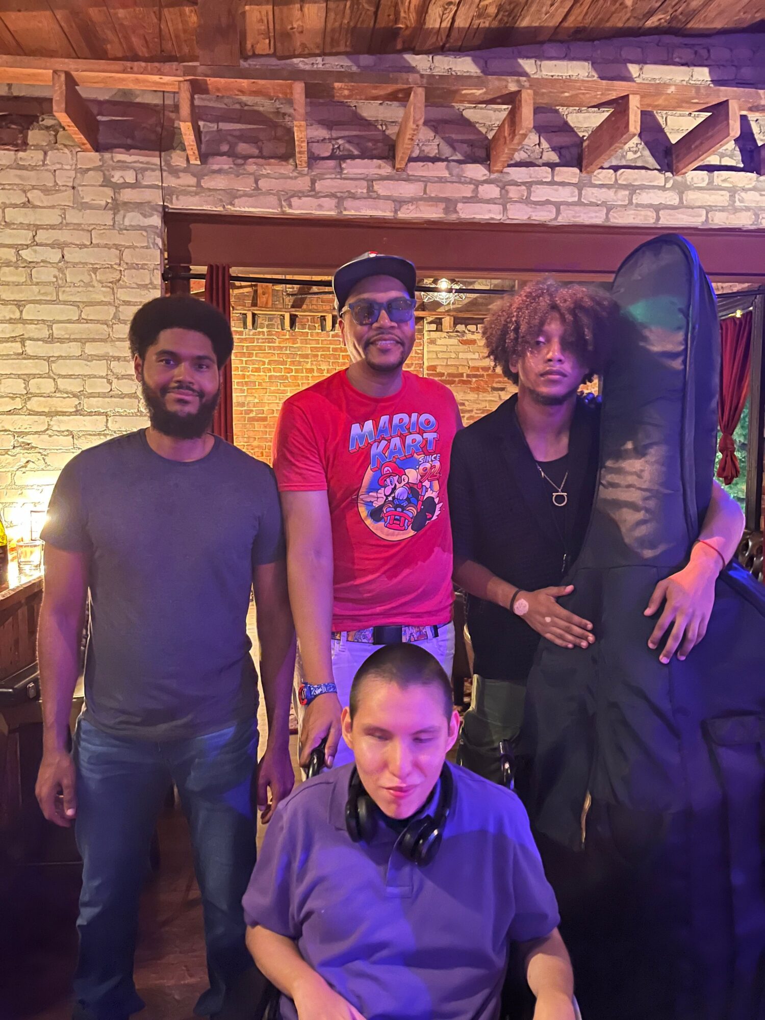 Four people standing indoors near a brick wall and wooden shelves, smiling and posing for the photo.