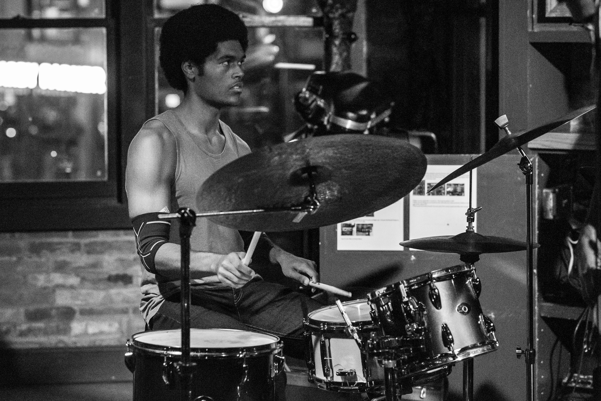A person playing drums in a room with large windows, wearing a sleeveless shirt and arm bands, in black and white.