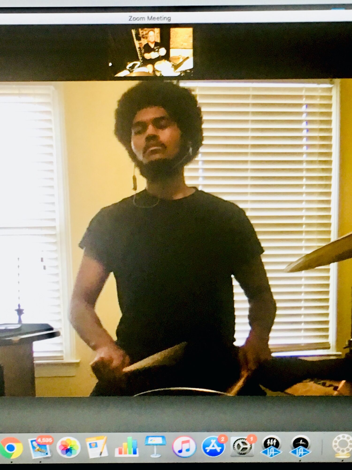 Person with curly hair holding drumsticks, standing indoors near window blinds, wearing a black shirt.