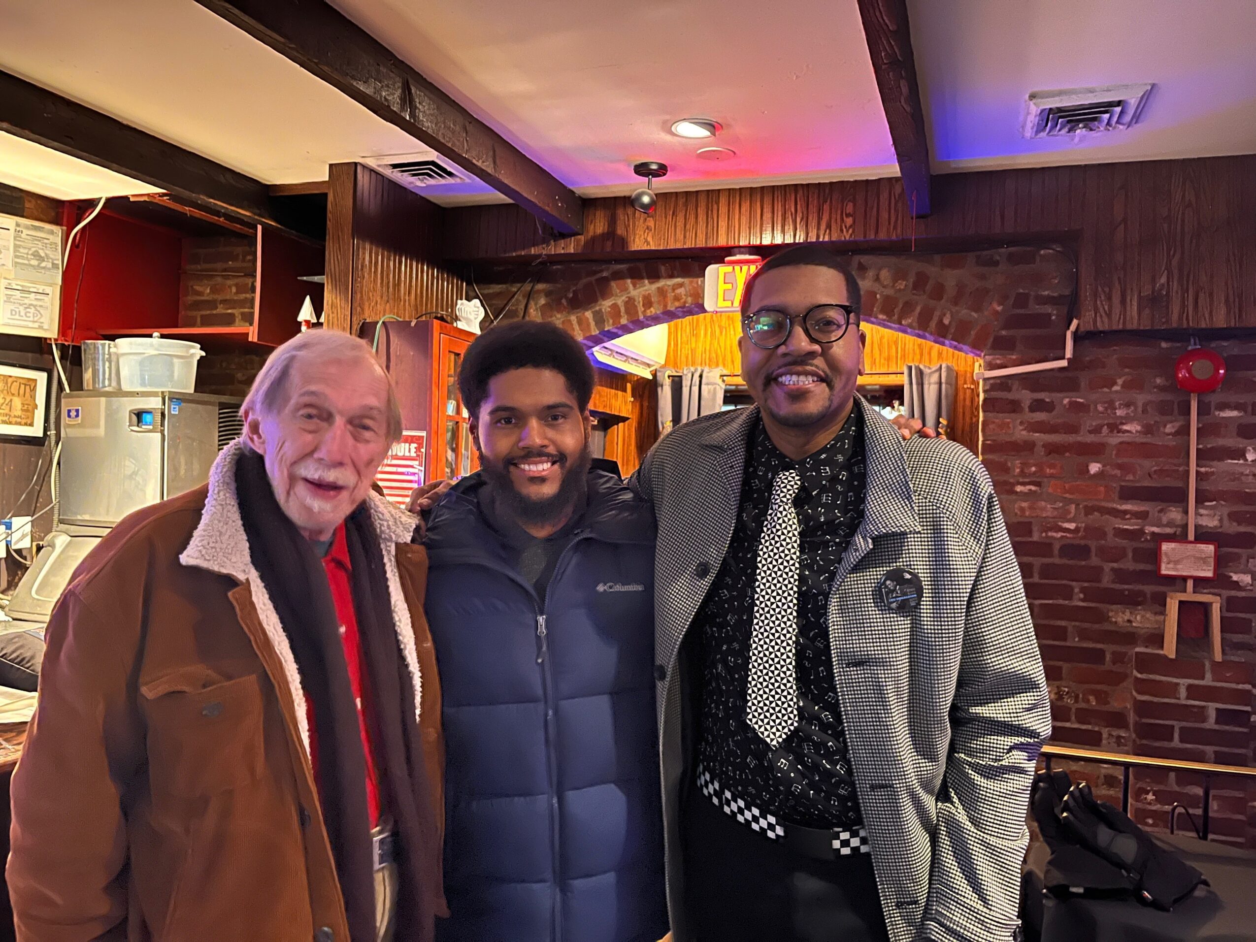 Three men standing together indoors, smiling, with brick wall and wooden decor in background.
