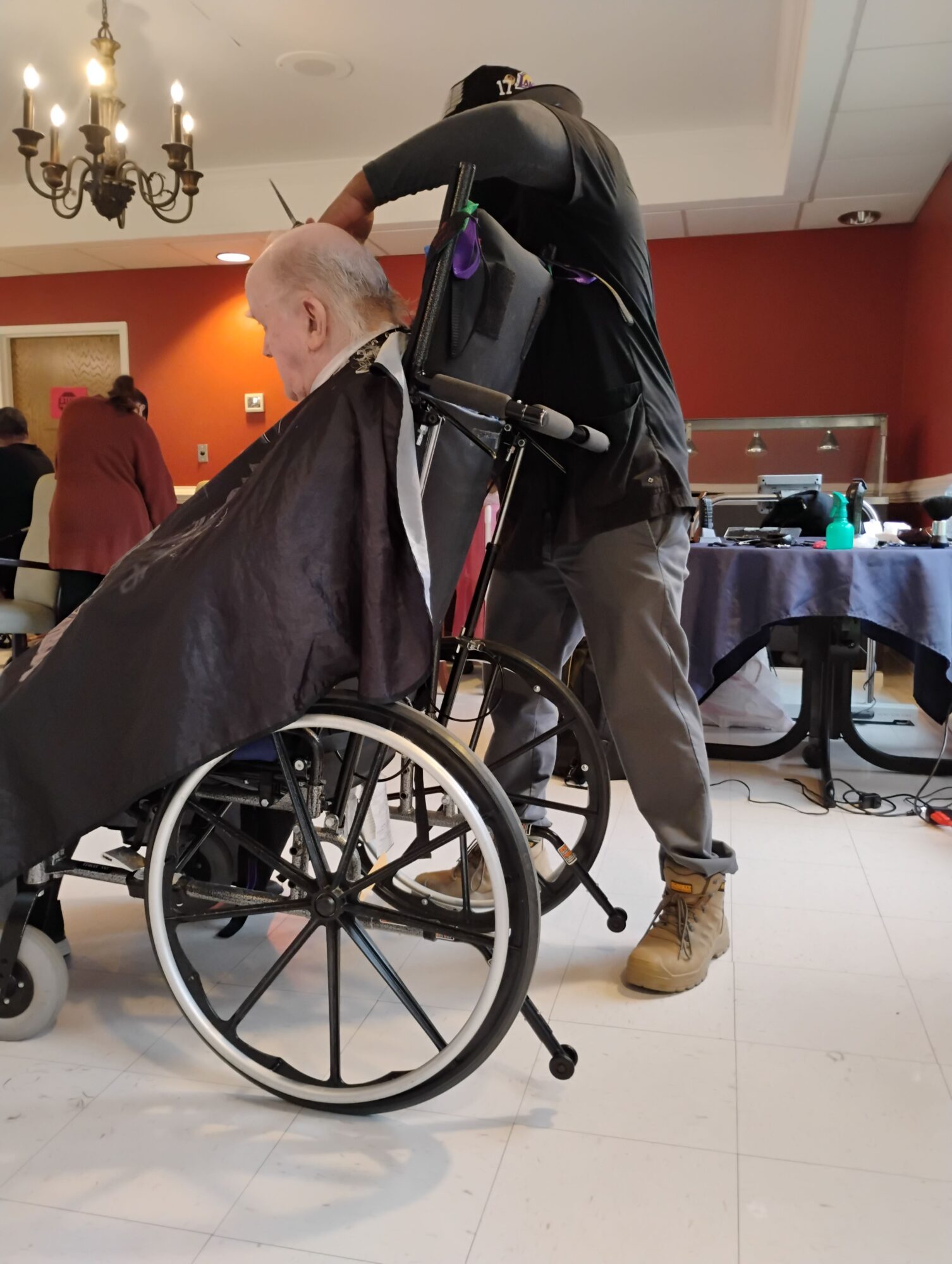 Person with short hair in a wheelchair, wearing a dark jacket, in an indoor setting with tables and chairs.
