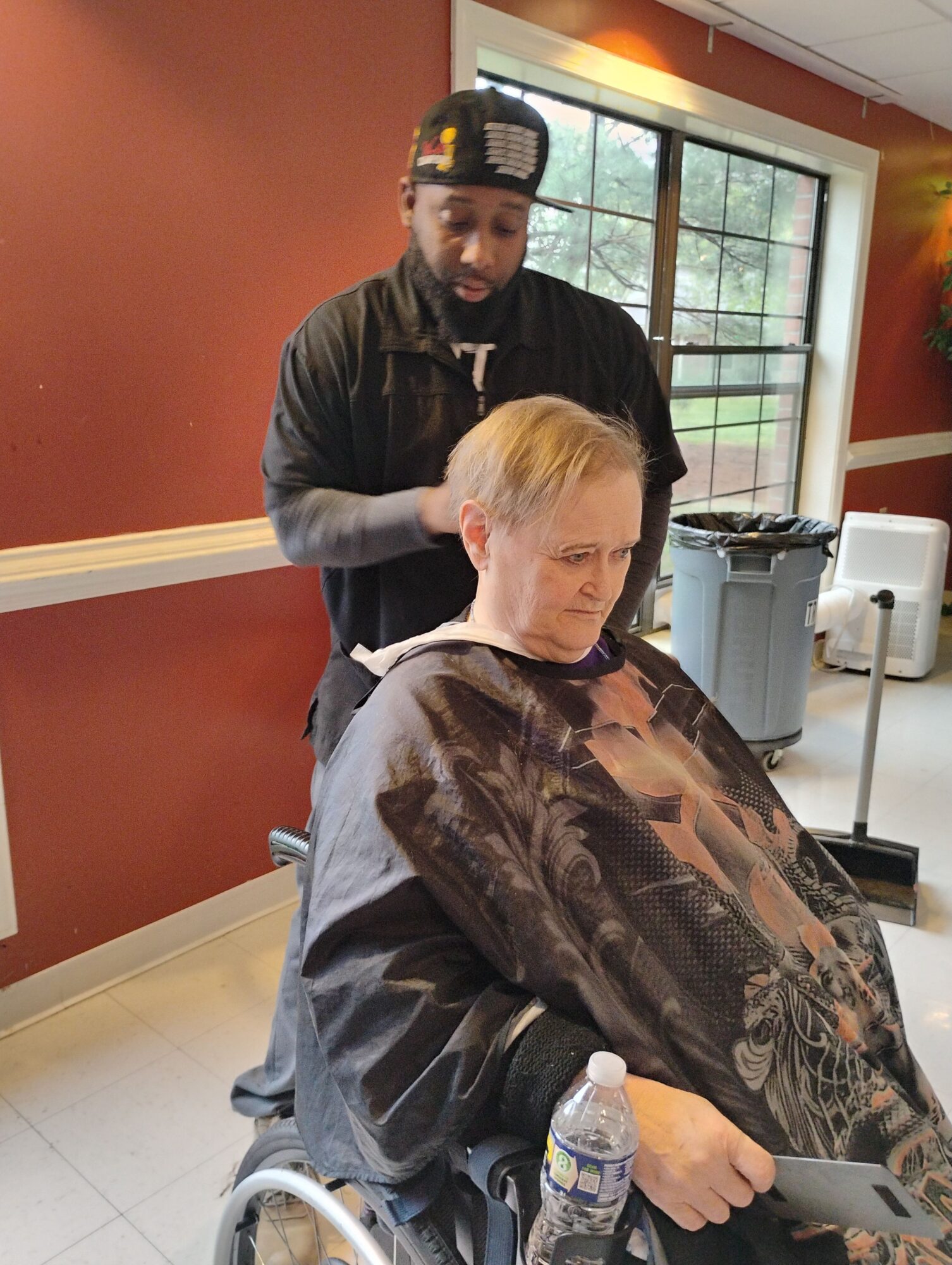 Person receiving a haircut in a salon, seated in a chair with a cape, with a stylist behind them, in a room with a window and red walls.