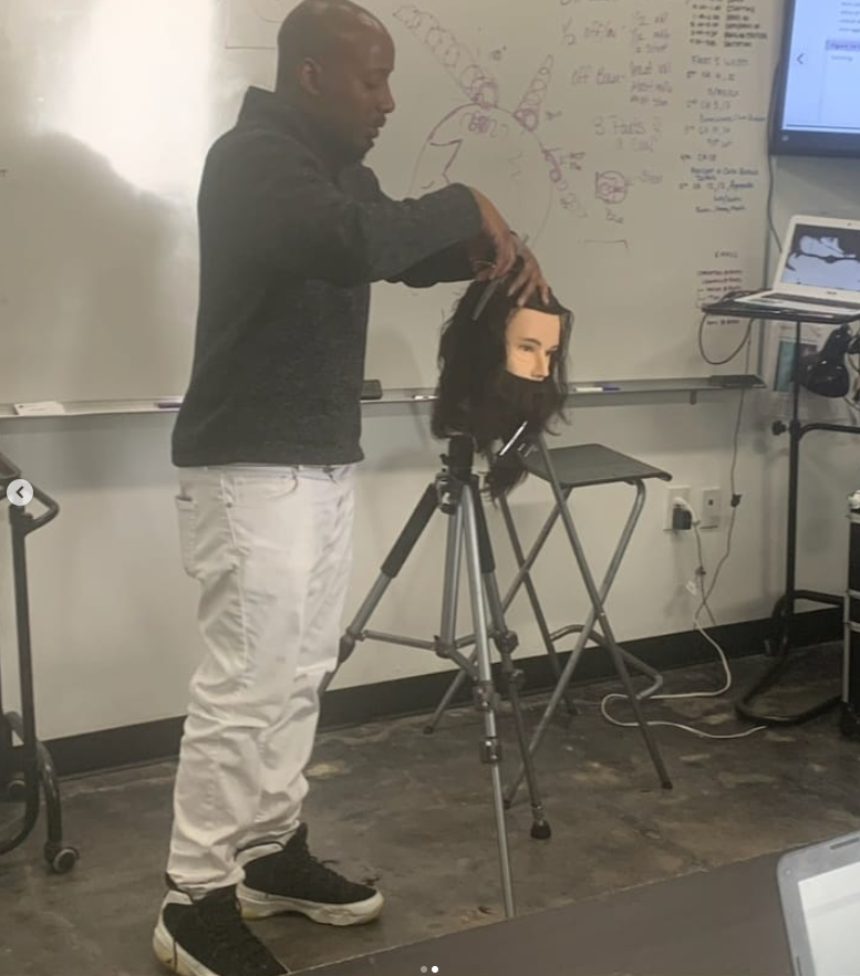 Person practicing hairstyling on mannequin head in classroom with whiteboard and monitor, standing next to a black chair.