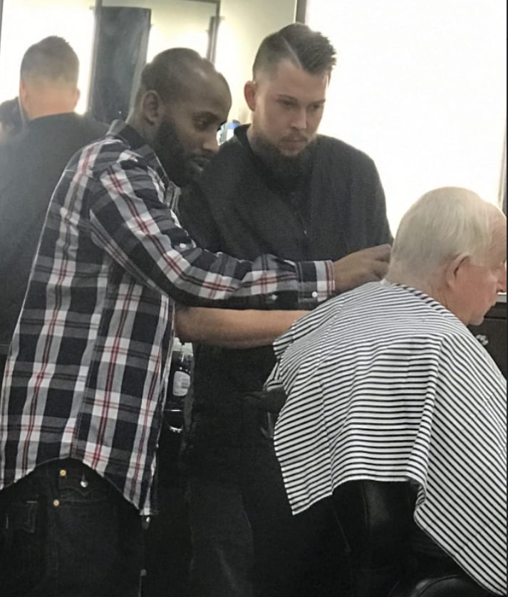 Two men standing behind a seated person with white hair, one in a plaid shirt and the other in black, in a salon. Word count: 20