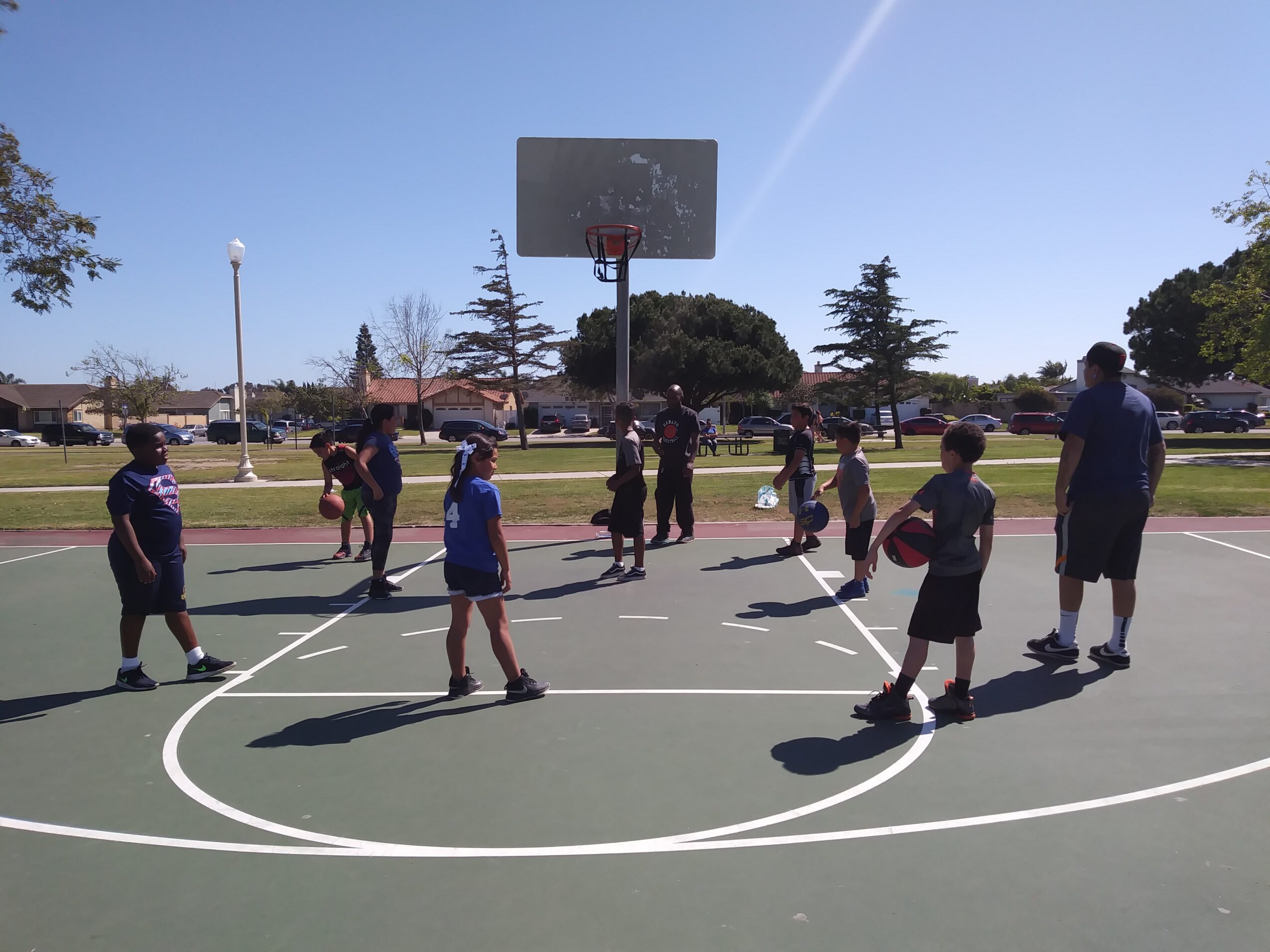 Group of children and an adult on a basketball court outdoors, with a basketball hoop and trees in the background.