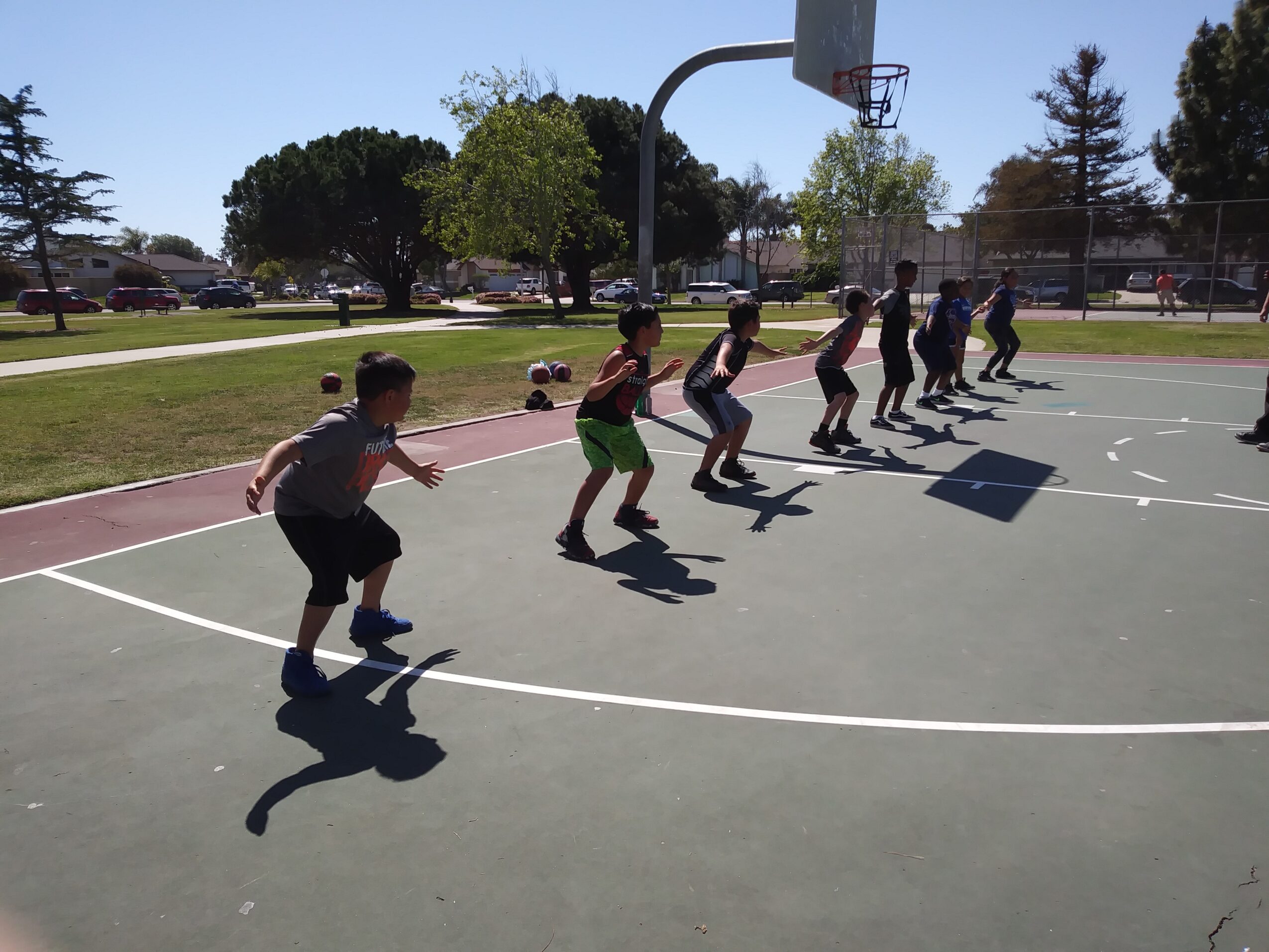 Children are playing basketball on an outdoor court, running and jumping in a line, with trees and parked cars in the background.