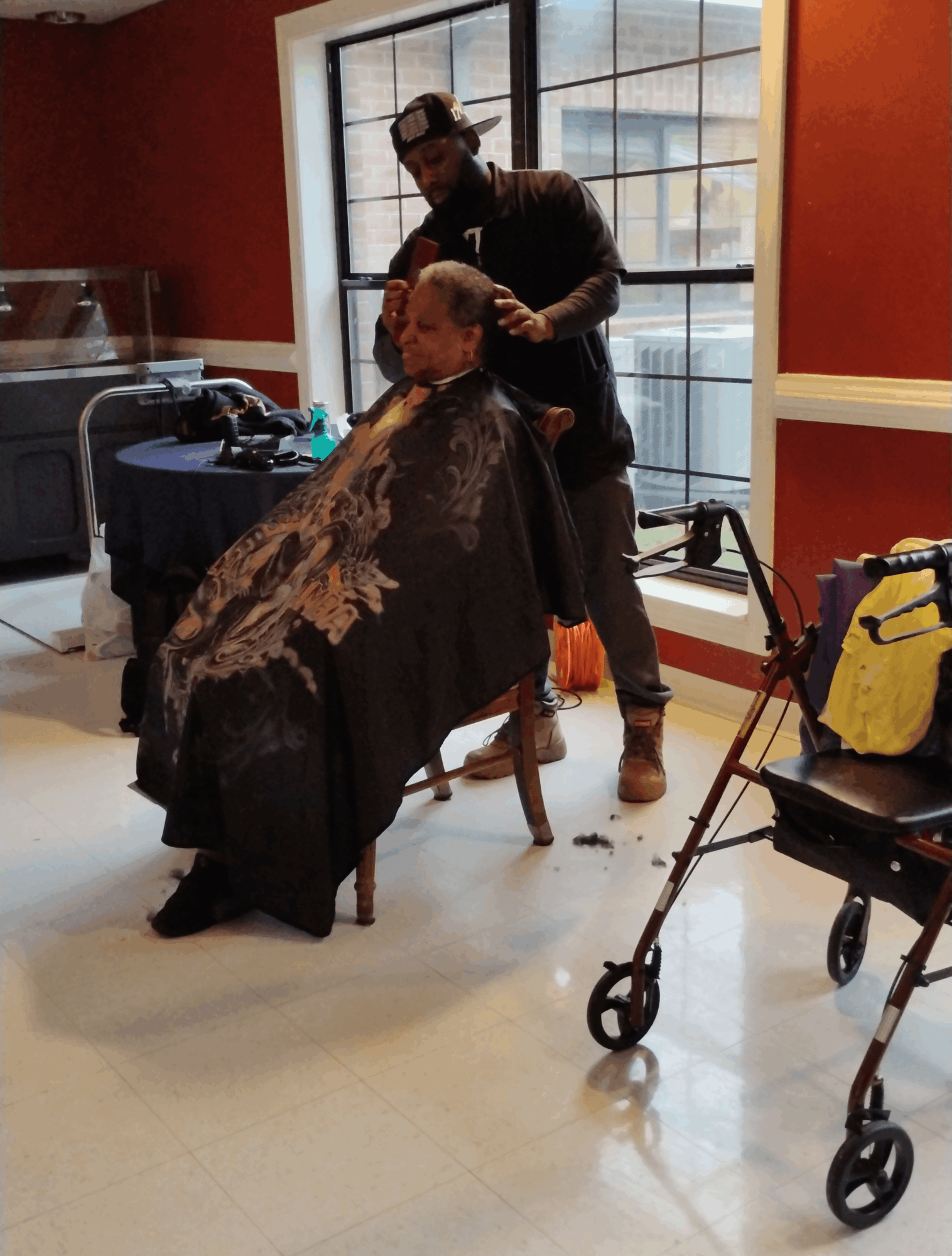 Person receiving a haircut in a salon with a stylist standing behind them, near a large window, with a walker nearby.