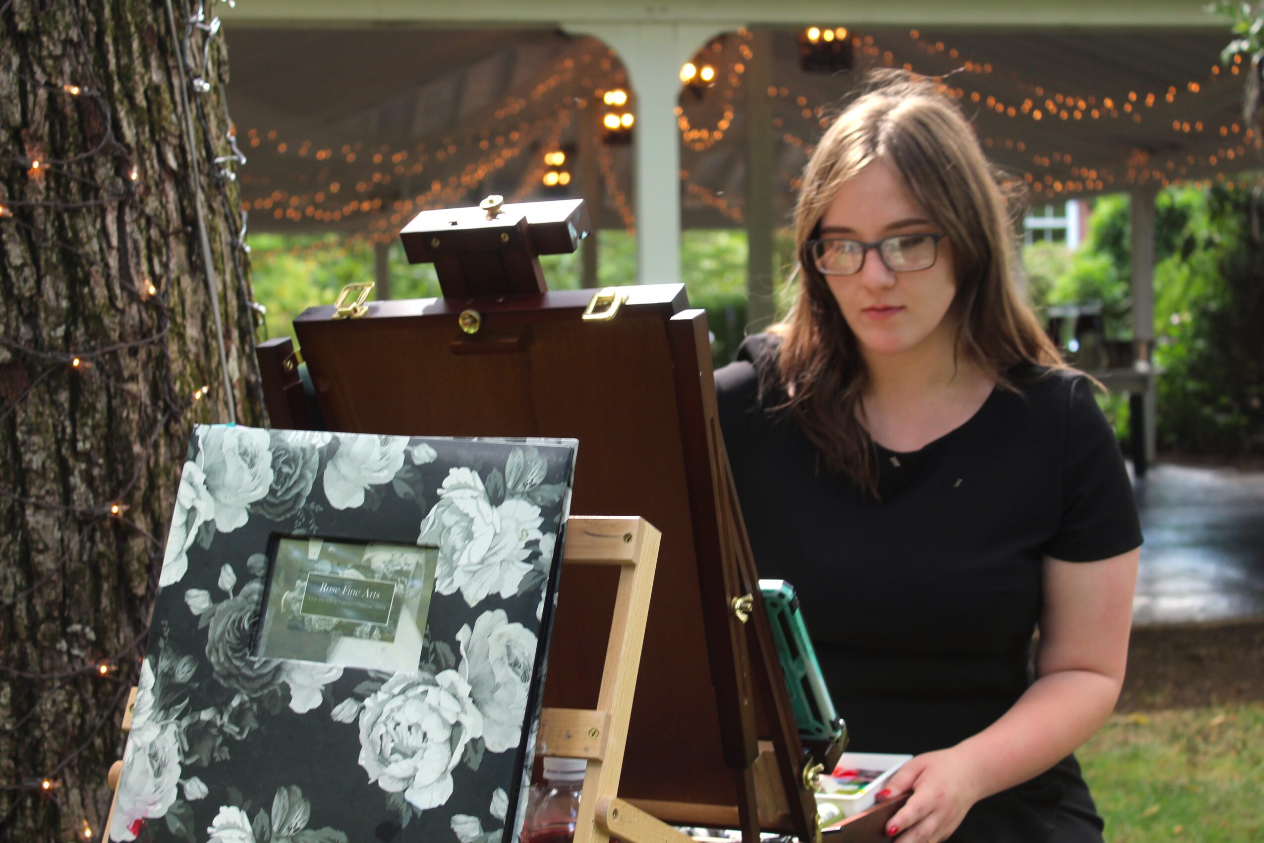 Young woman with glasses and long hair, wearing a black shirt, standing outdoors near a tree with a display board.