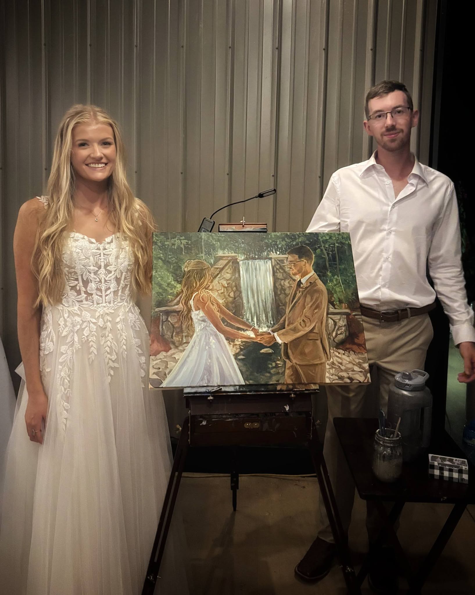 A woman in a white dress and a man in a white shirt stand beside a painting of a wedding scene, with a curtain background.