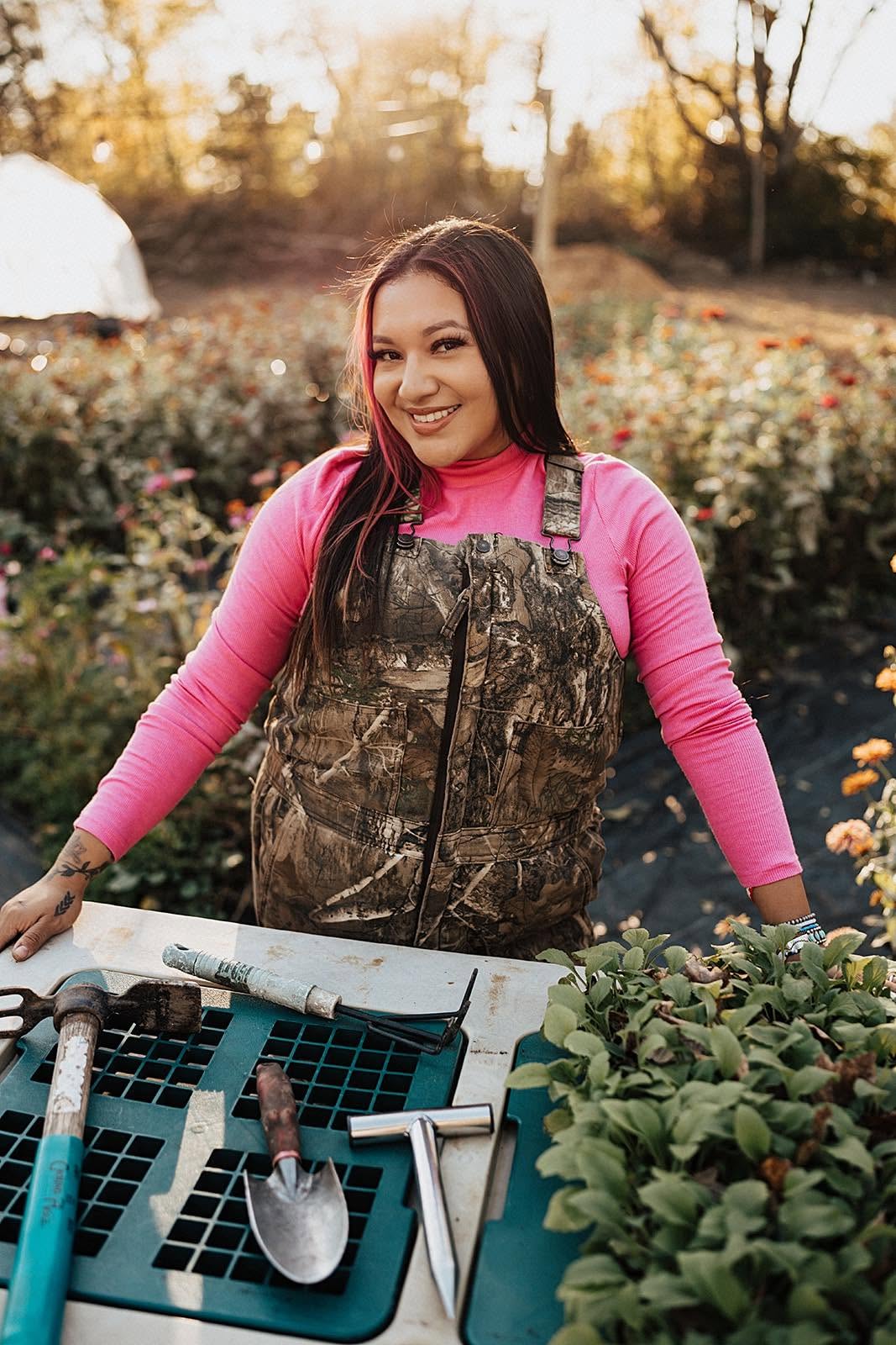 Young woman in camouflage apron and pink shirt smiling outdoors among plants and flowers.