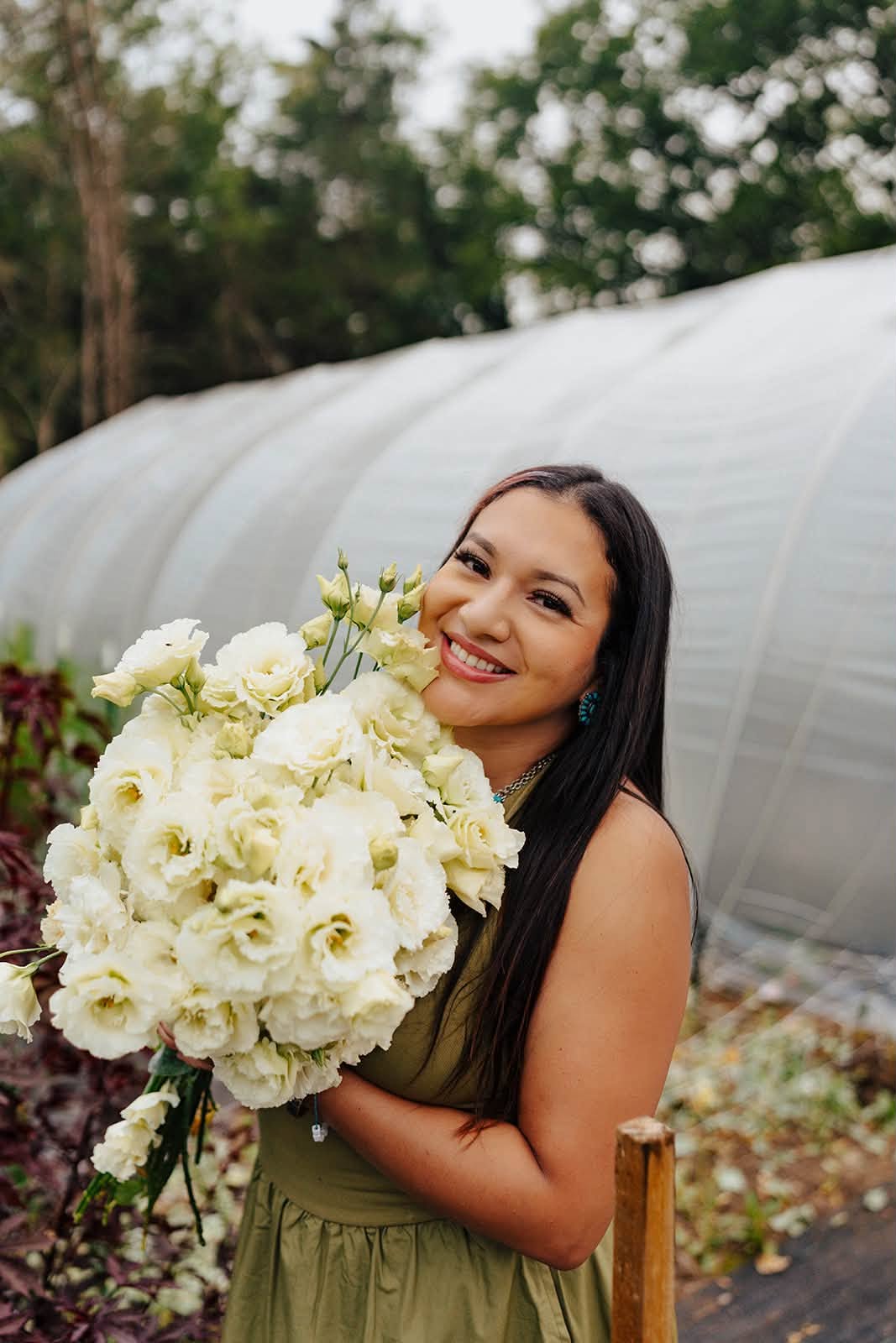 Woman holding large bouquet of white flowers outdoors, smiling, with greenery and a greenhouse in background.