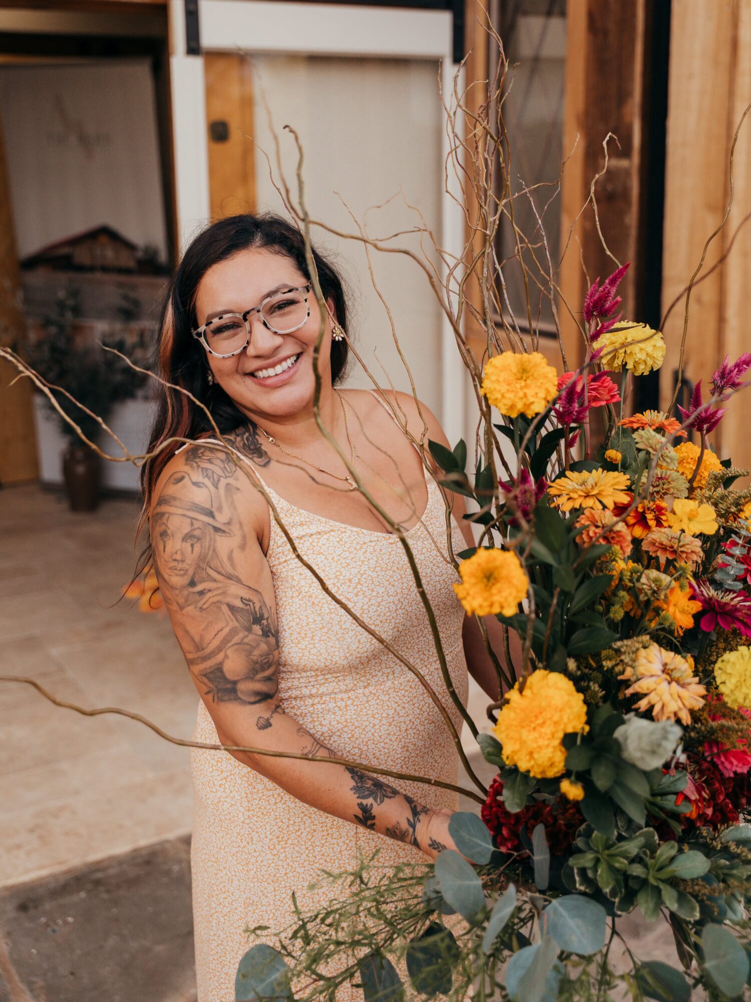 Woman with glasses and tattoos smiling next to a colorful flower arrangement indoors.