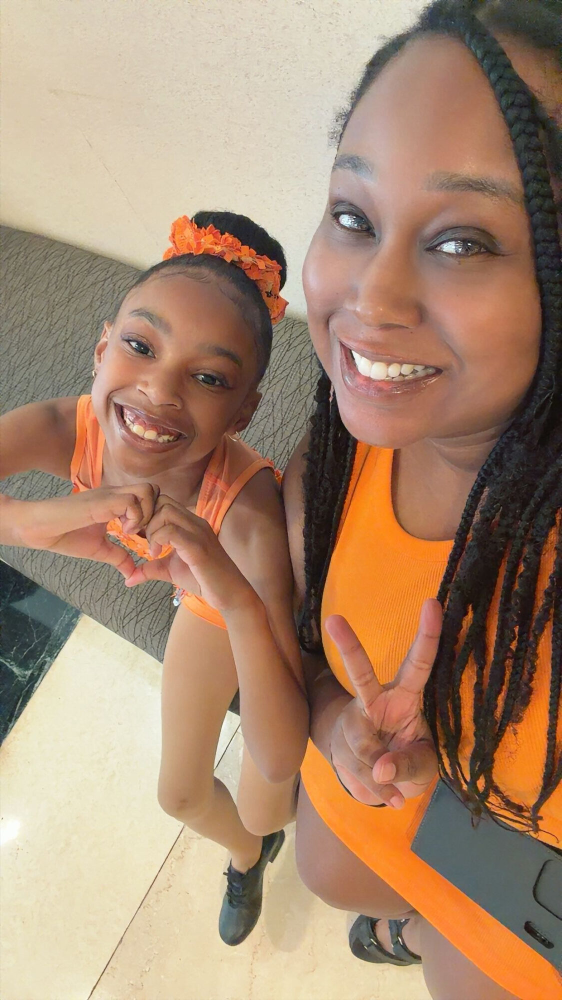 Two smiling females, one adult and one child, taking a selfie together, both wearing orange outfits, indoors.