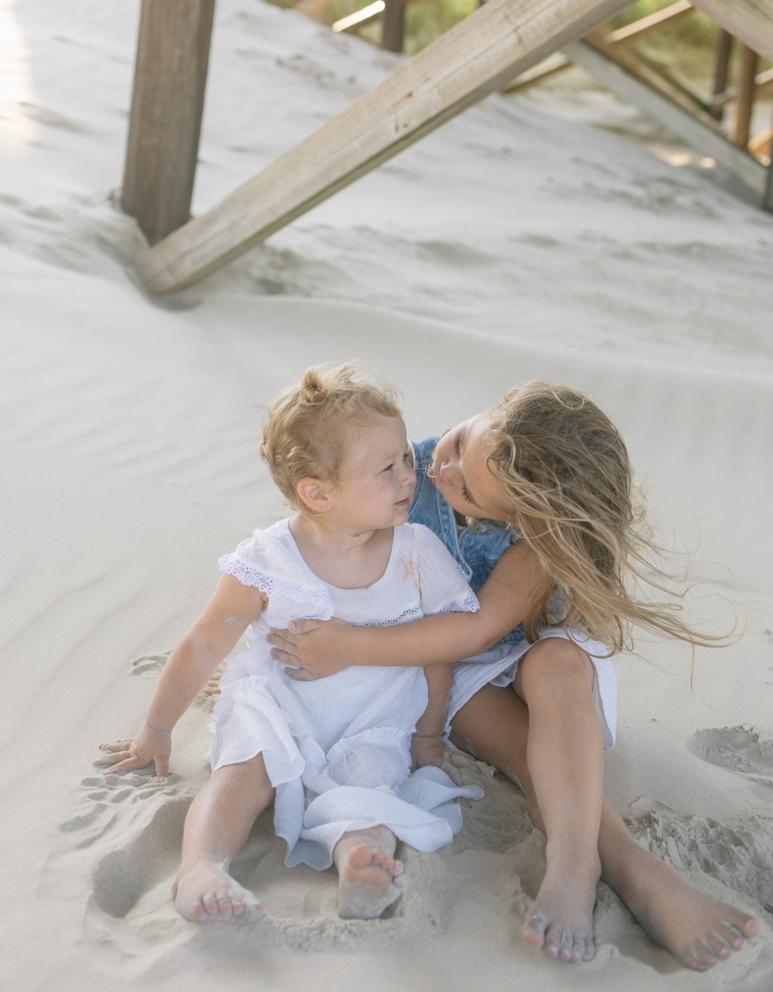 Two children sitting on sand at the beach, one with long hair and the other with short hair, embracing each other.