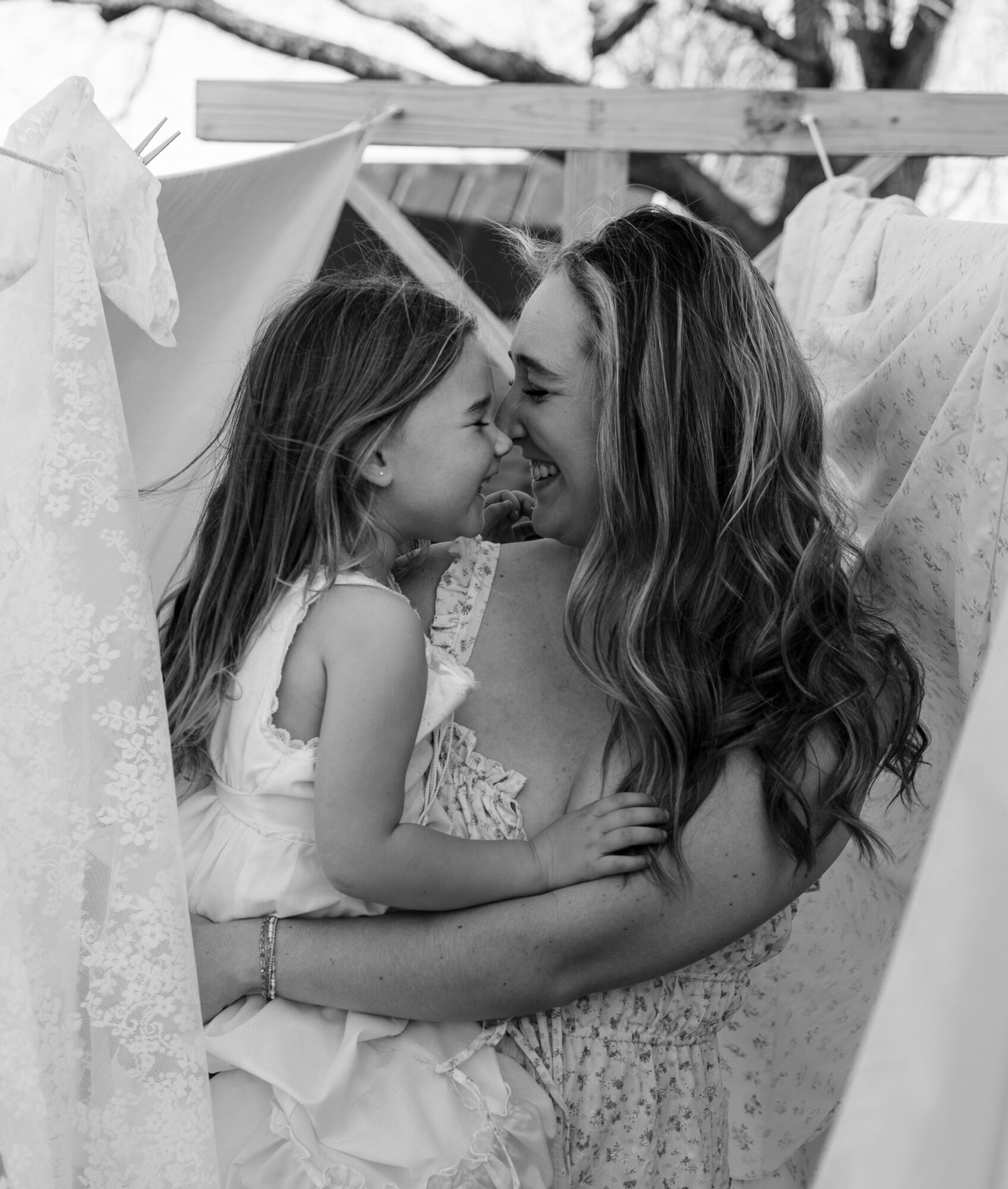 Woman and young girl face each other smiling, surrounded by hanging laundry outdoors.