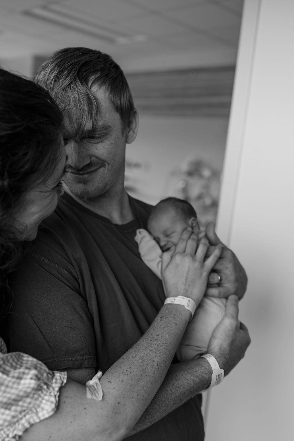 Man holding a newborn baby, with a woman leaning in, in a hospital room, black and white photo.
