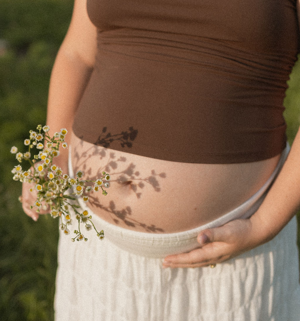 Pregnant woman holding flowers on her belly outdoors, shadow of flowers on her stomach, wearing a brown top and white skirt.