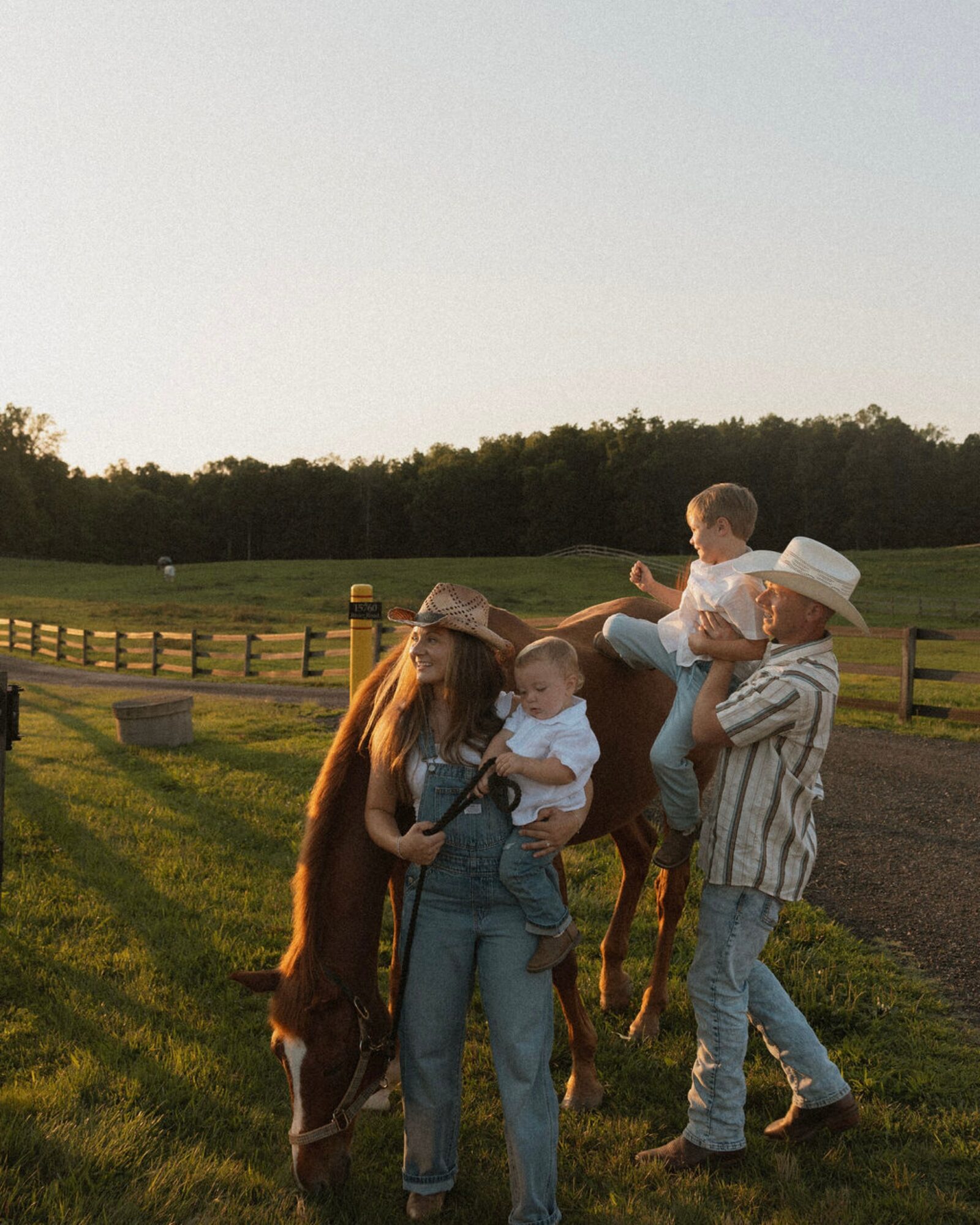 Group of people outdoors with a horse, children, and adults, standing on a grassy area near a wooden fence, under a clear sky.