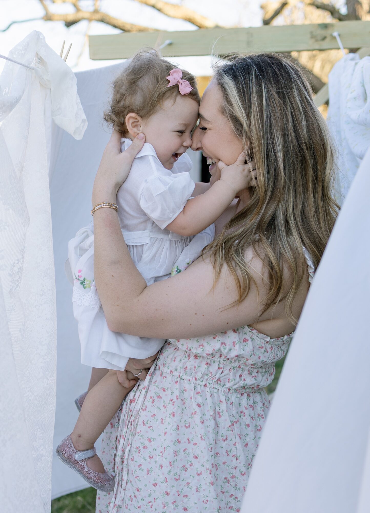 Woman holding a young girl close, both smiling and touching foreheads outdoors, with laundry hanging in the background.