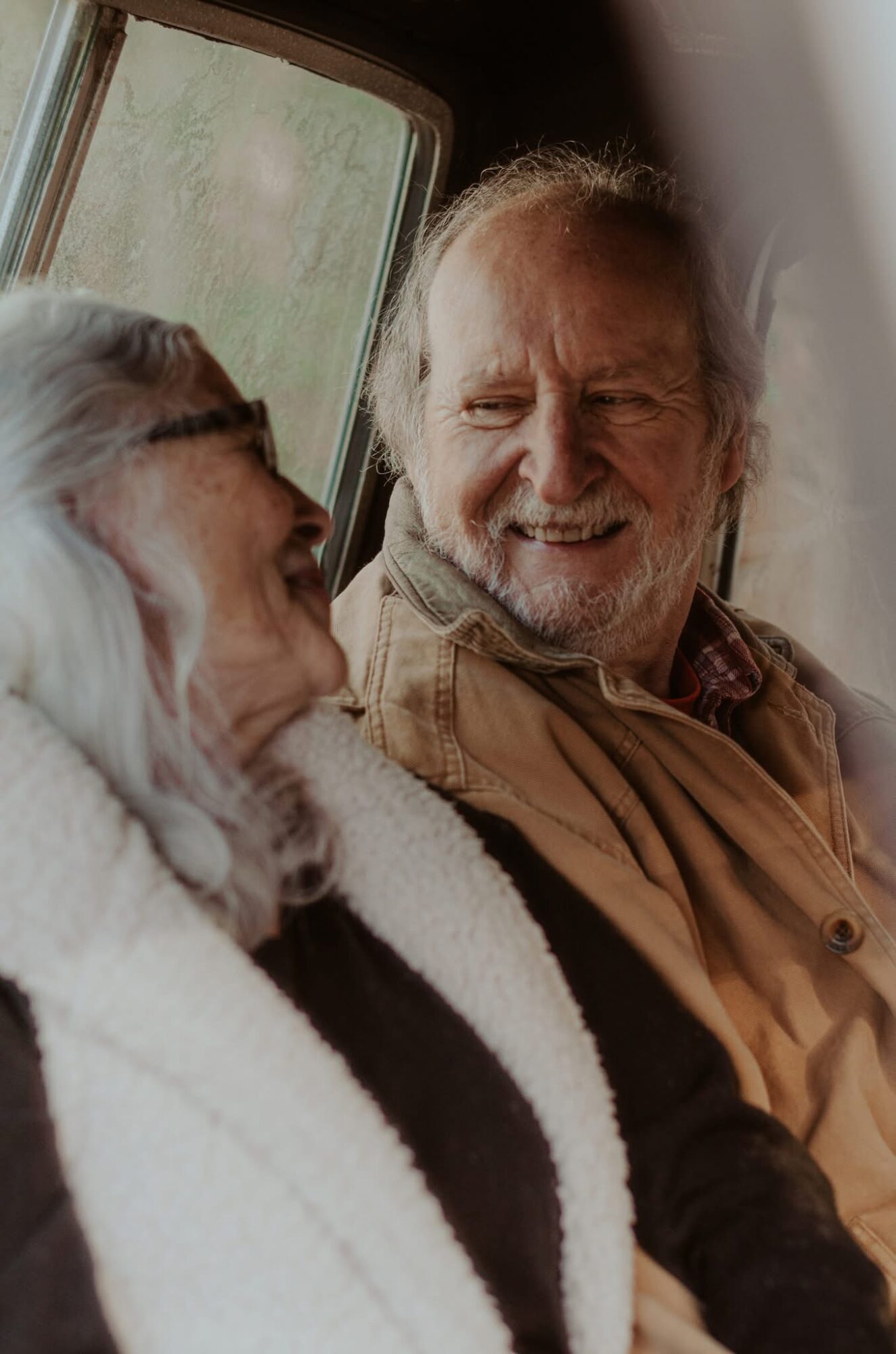 Two elderly people smiling and looking at each other inside a vehicle, with a window showing greenery outside.