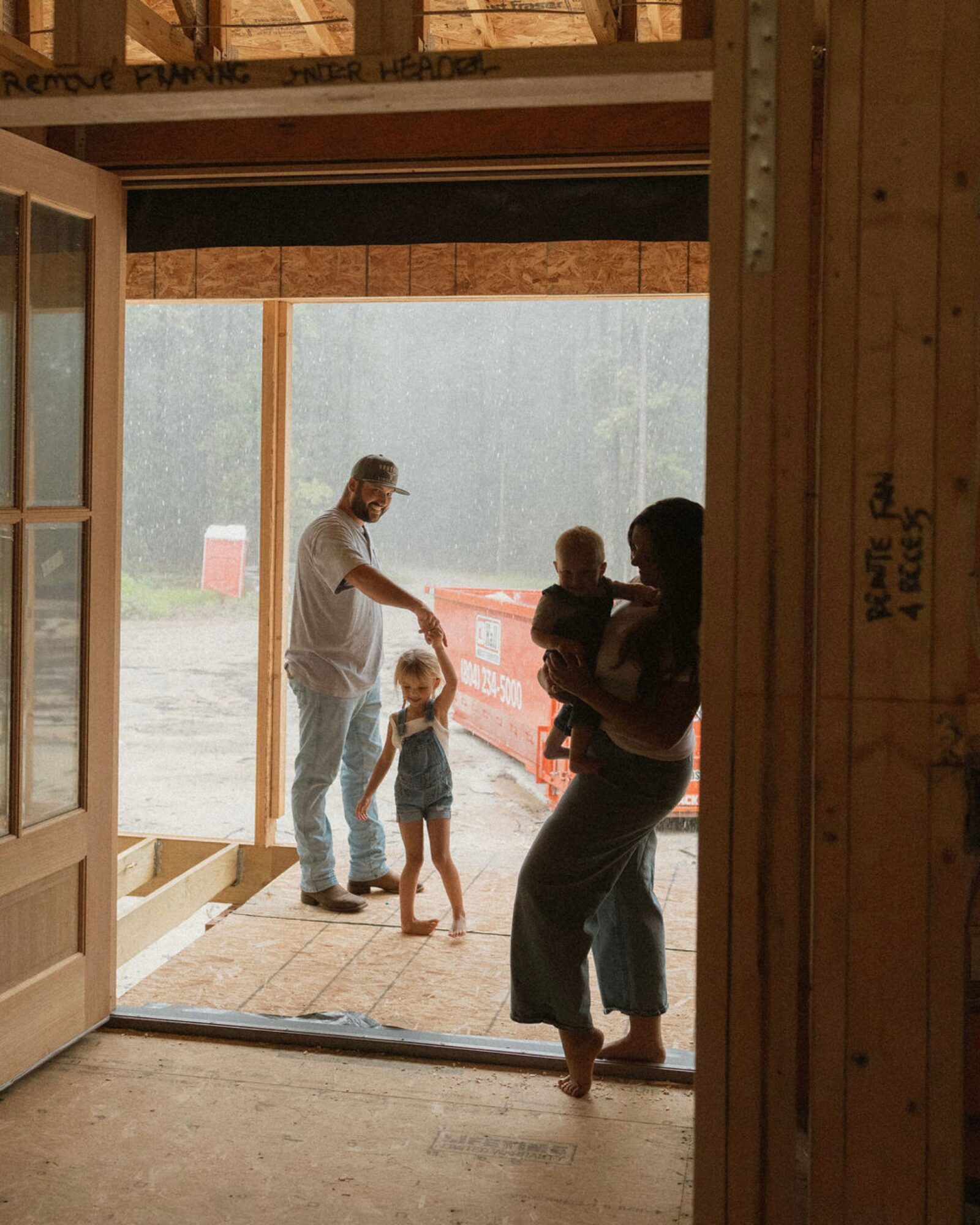 People standing at an open doorway, with a person holding a child, outside view of trees and fog, wooden interior.