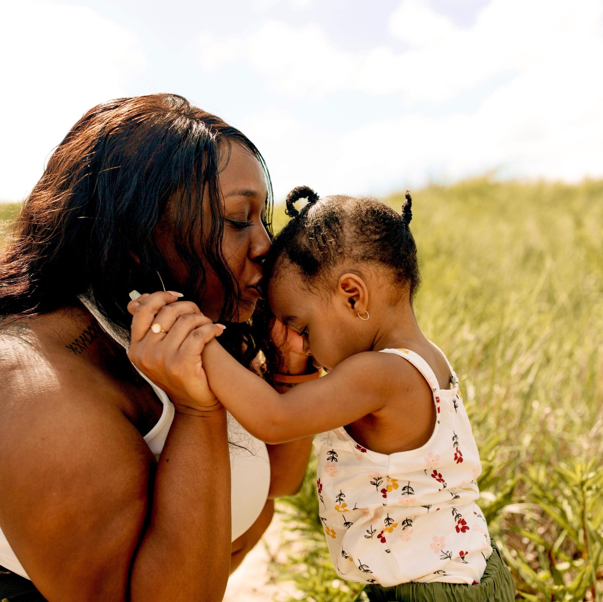 Woman and young girl touching foreheads outdoors in a grassy field, sunlight in background.