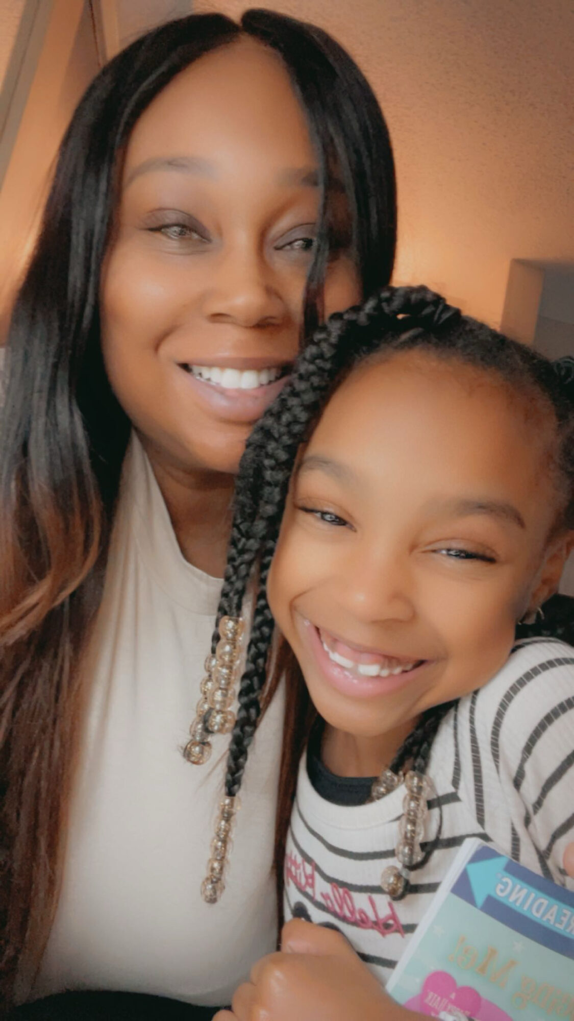 Two smiling females, one adult and one child, close together indoors, both with braided hair, smiling at the camera.