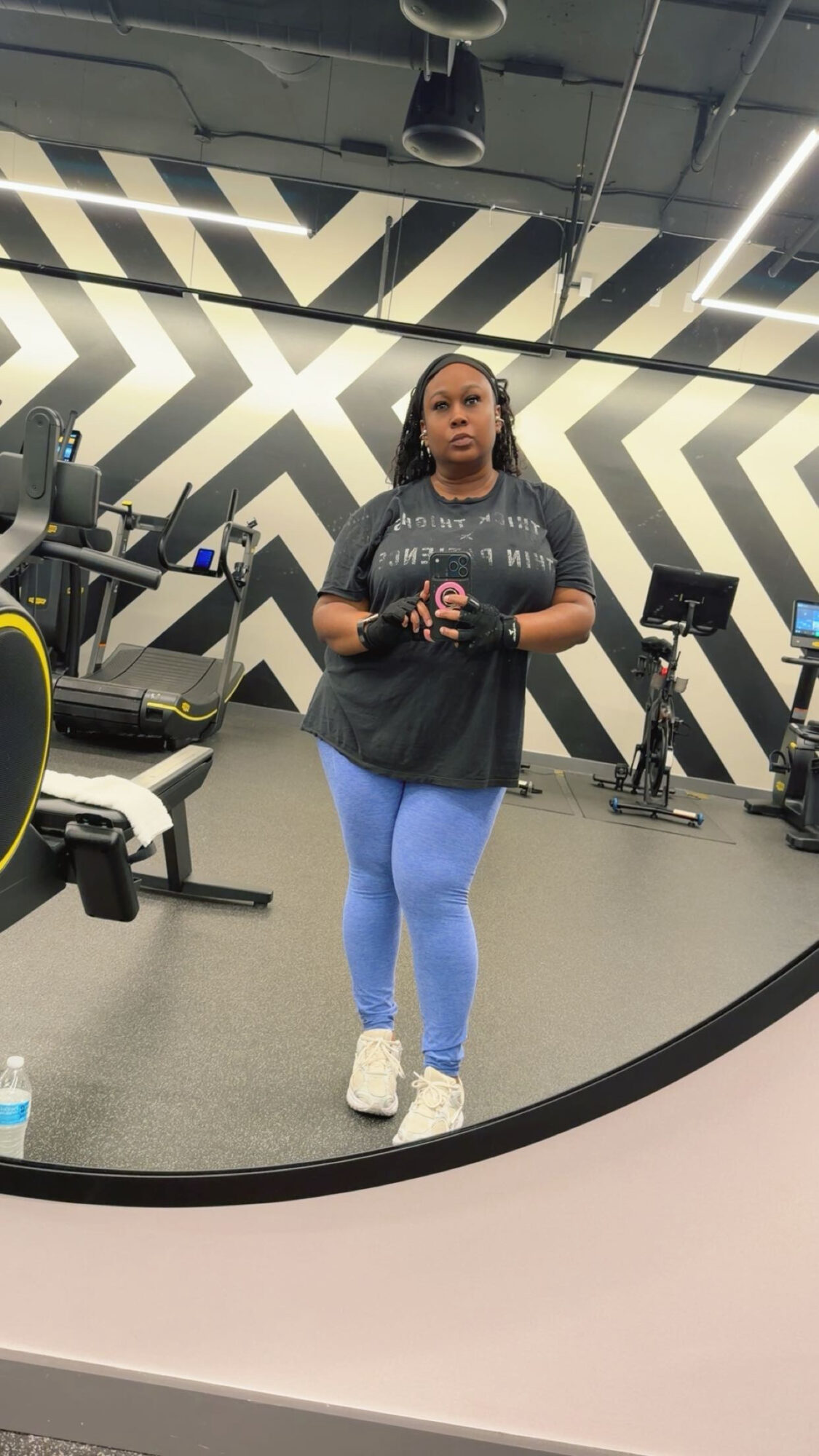 Woman taking a mirror selfie in a gym, wearing a black top, blue leggings, and white shoes, with gym equipment in background.