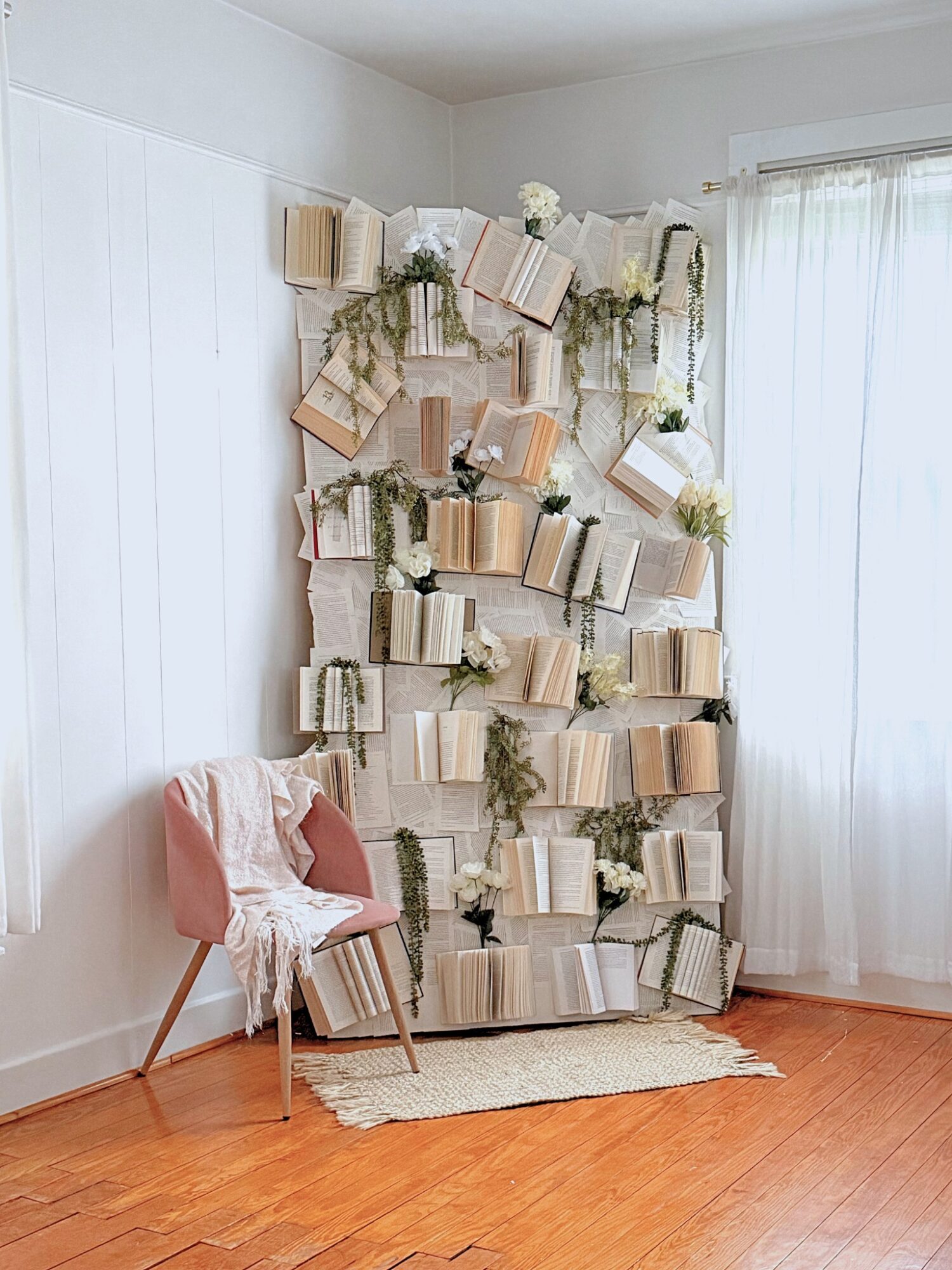 Books arranged vertically on a wall with greenery, next to a pink chair with a blanket, in a bright room.