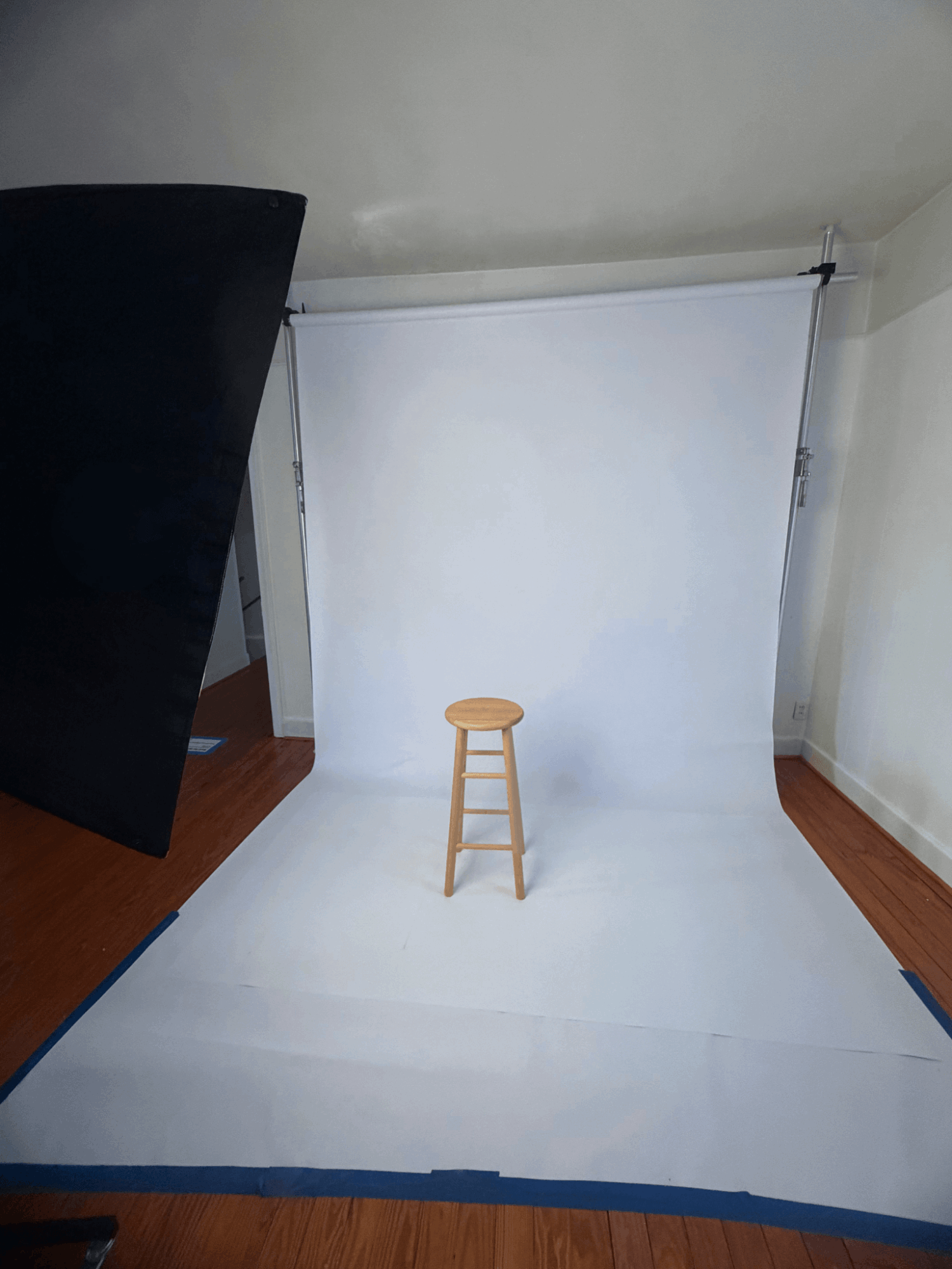 White backdrop with a wooden stool in front, set up in a room with hardwood floor and white walls.