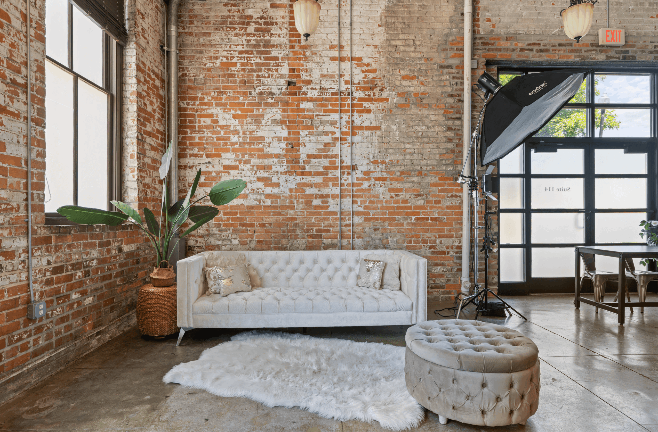 Interior with brick wall, white sofa, potted plant, round ottoman, and large window with natural light.