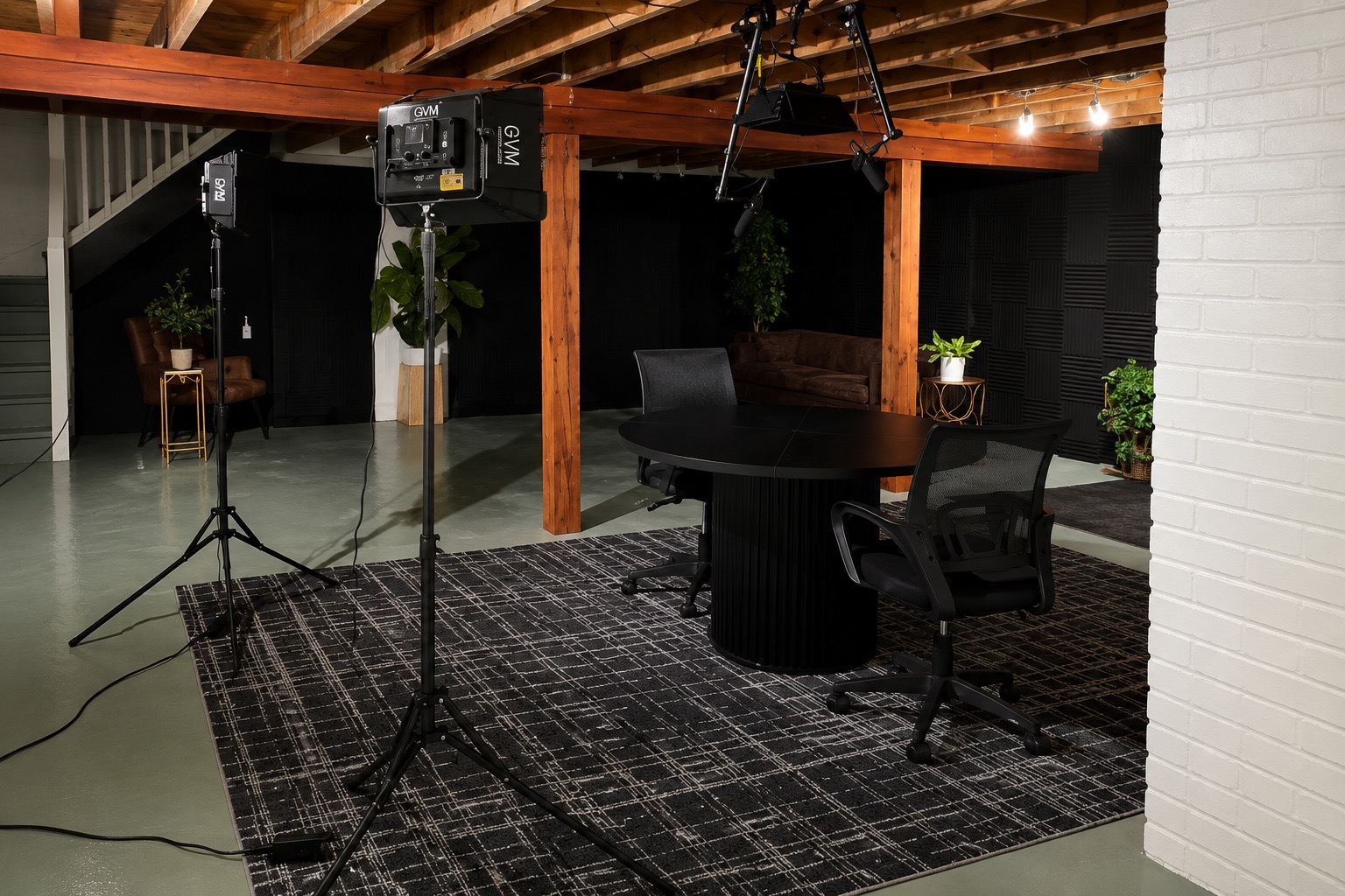 Indoor space with wooden beams, a round table, chairs, potted plants, and professional lighting equipment on a patterned rug.