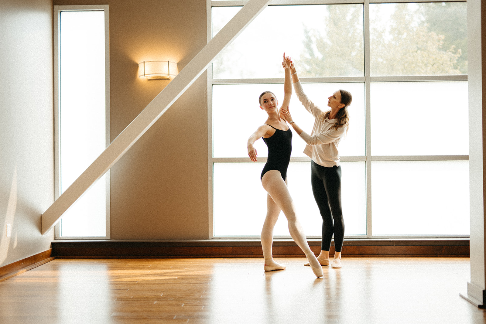 Two women practicing ballet in a dance studio with large windows, one holding a pose while the other assists.