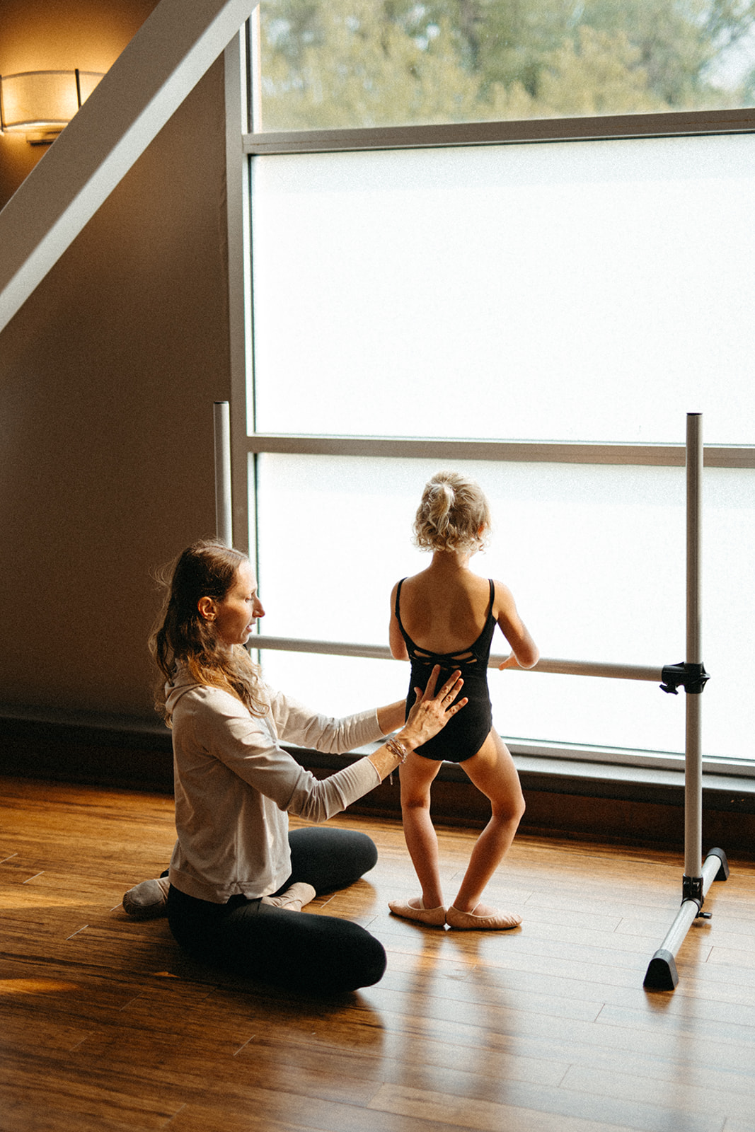 Adult woman assisting a young girl with ballet practice near a large window, wooden floor, and ballet barre, daytime.