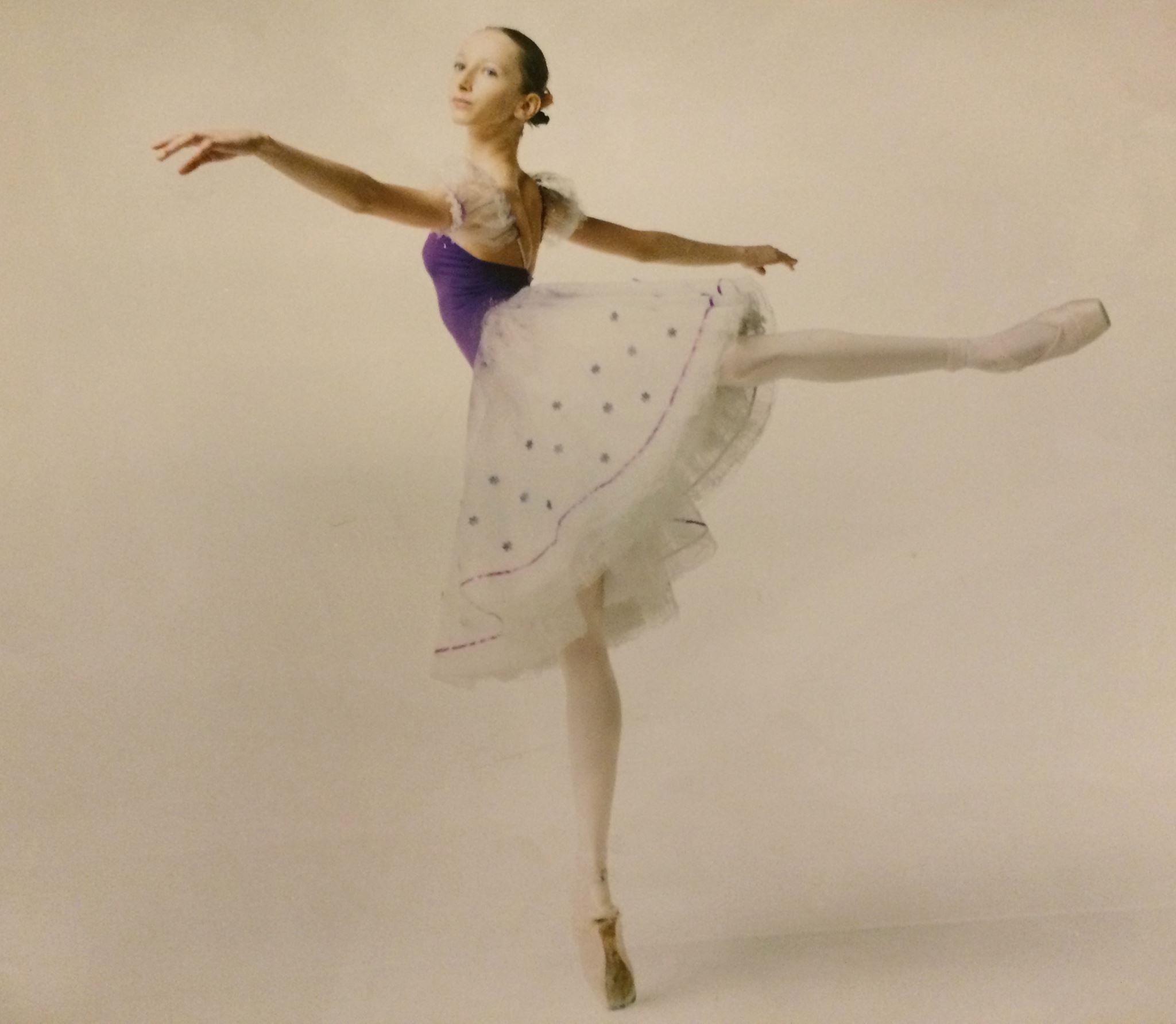 Ballet dancer in a purple and white tutu performing a pose with one leg extended behind and arms outstretched.