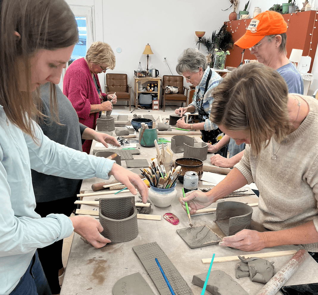 People working on pottery projects at a table in a classroom, with tools and clay. Six individuals are visible, focused on their work.