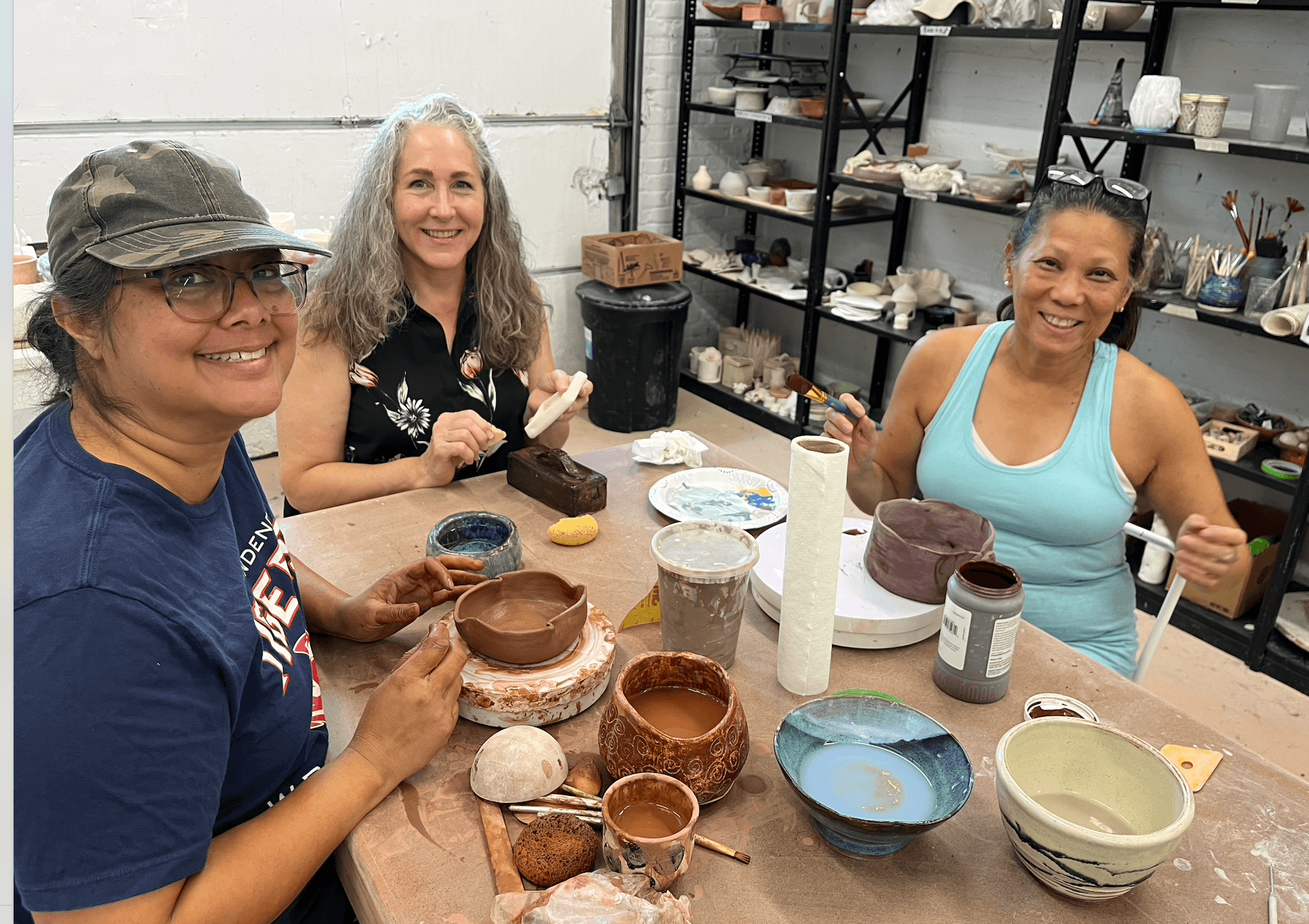 Three women sit at a table in a pottery studio, smiling with finished and in-progress clay pottery pieces.