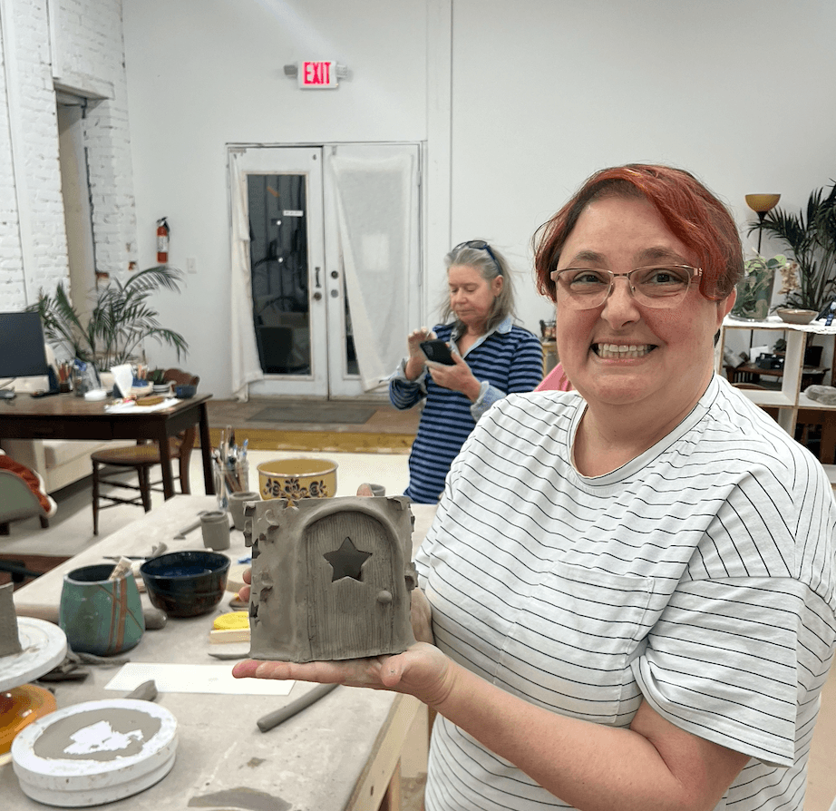 Woman smiling and holding a ceramic piece in a workshop, with another person in the background using a phone.