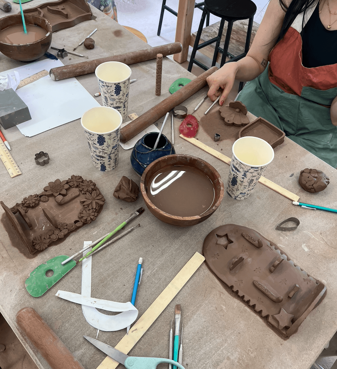 People are working on clay pottery projects at a table with tools, cups, and clay pieces, some shaped like vehicles.