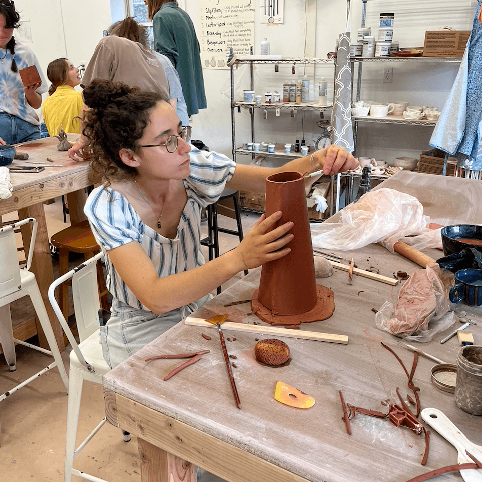Girl with glasses and curly hair working on a pottery project at a workshop table, surrounded by tools and clay.