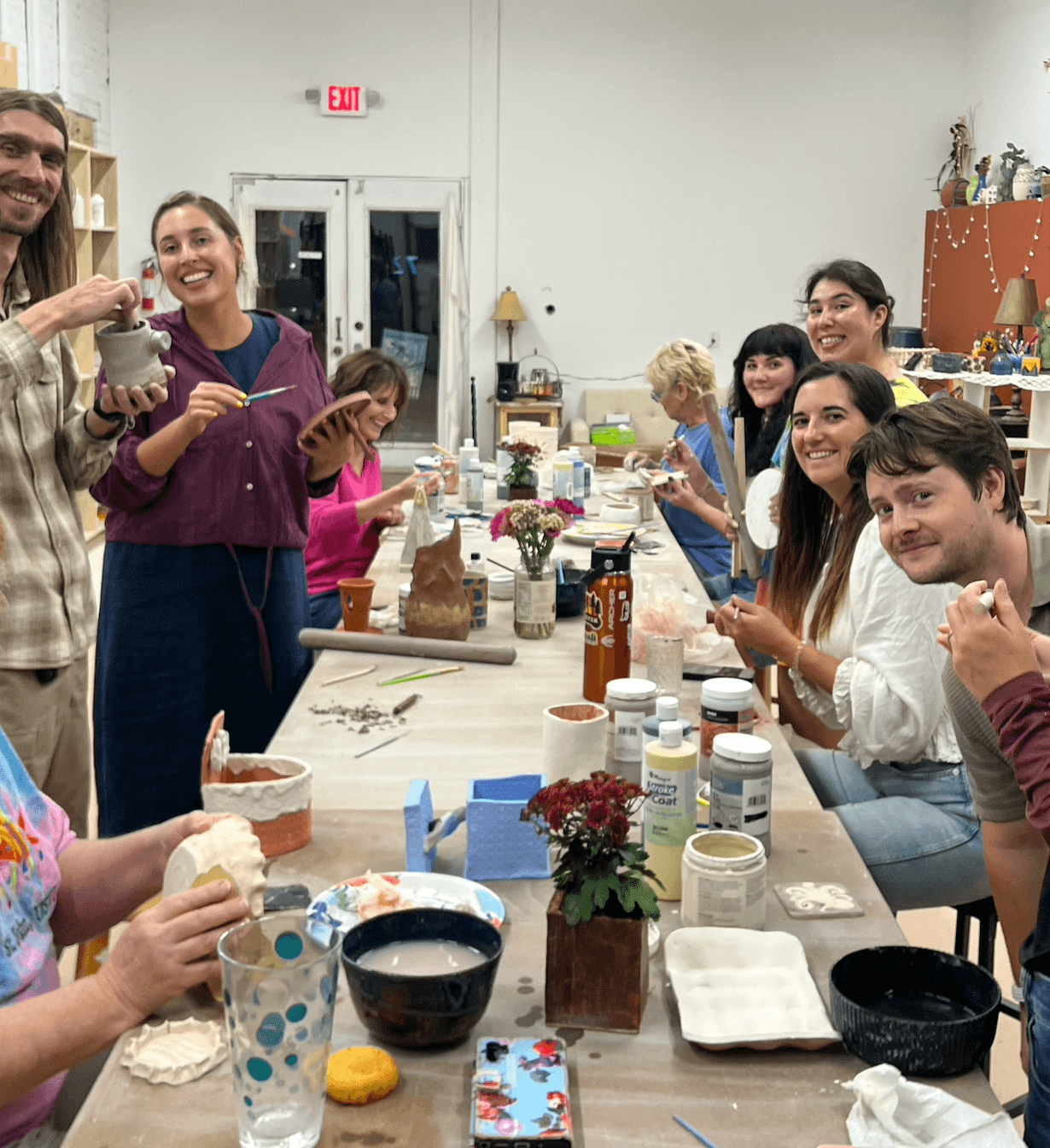 Group of people around a table with food and drinks, smiling and holding items, in a bright room with shelves and decorations.