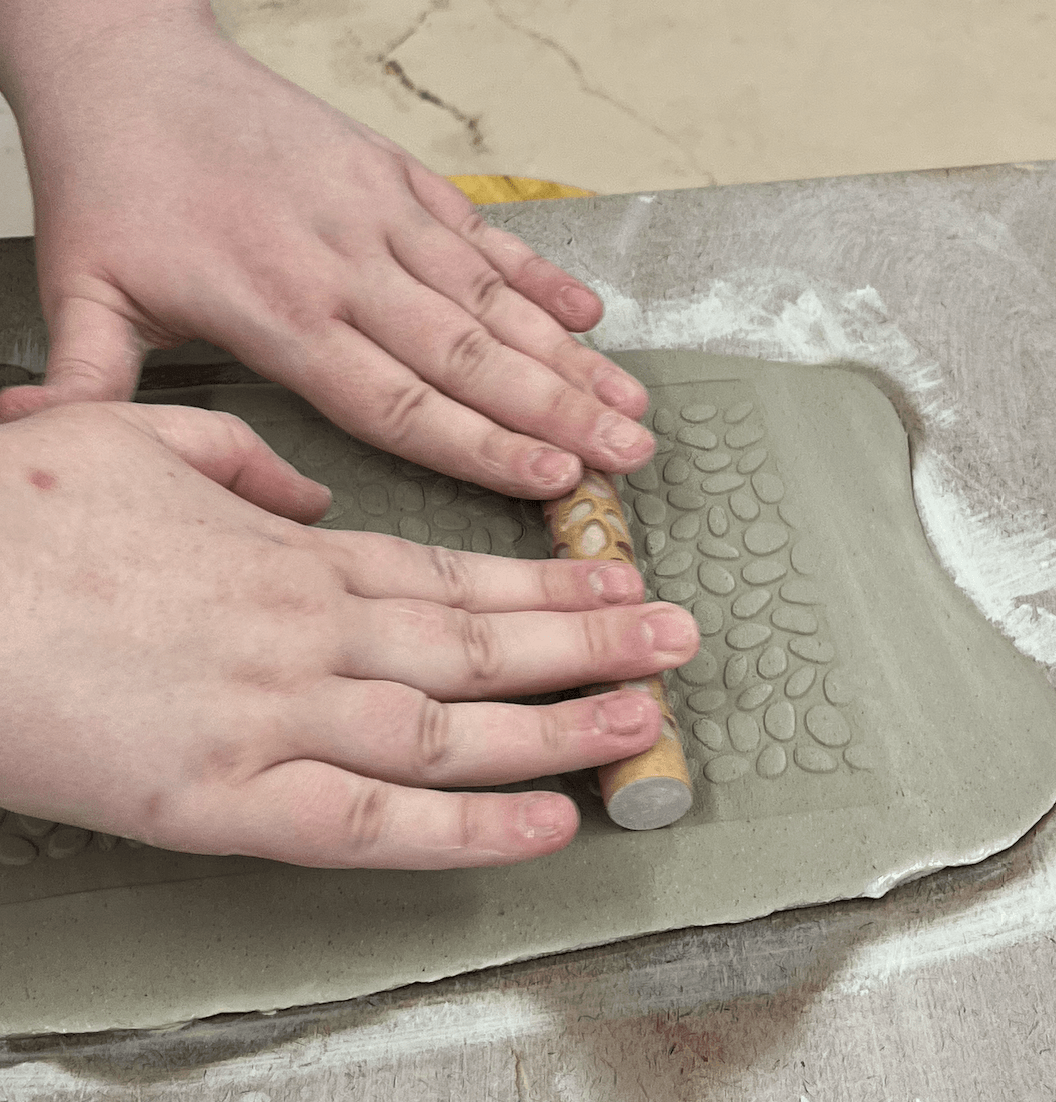 Hands pressing clay with a textured rolling pin on a work surface, dusted with flour, in a baking or pottery setting.