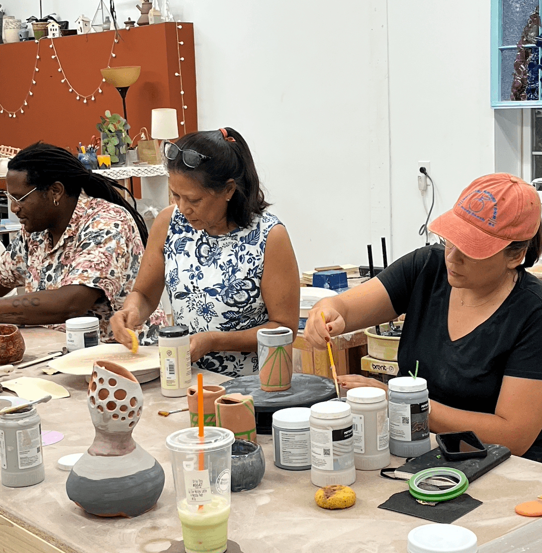 Three women sit at a table working on art projects with jars, brushes, and art supplies, in a bright room.