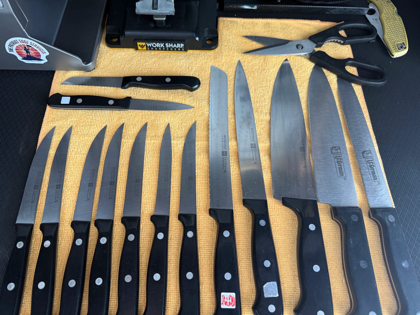 A collection of kitchen knives and scissors arranged on a yellow cutting mat, with a black box and a yellow utility knife nearby.