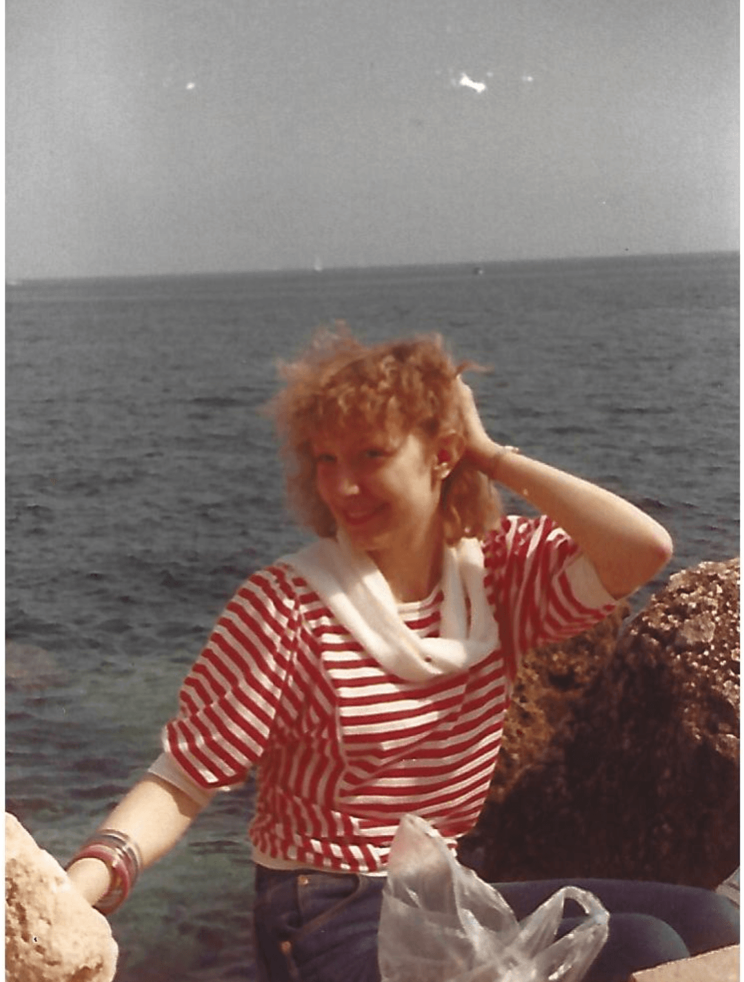Person with curly hair wearing a red and white striped shirt sitting by the water, smiling, with rocks nearby.