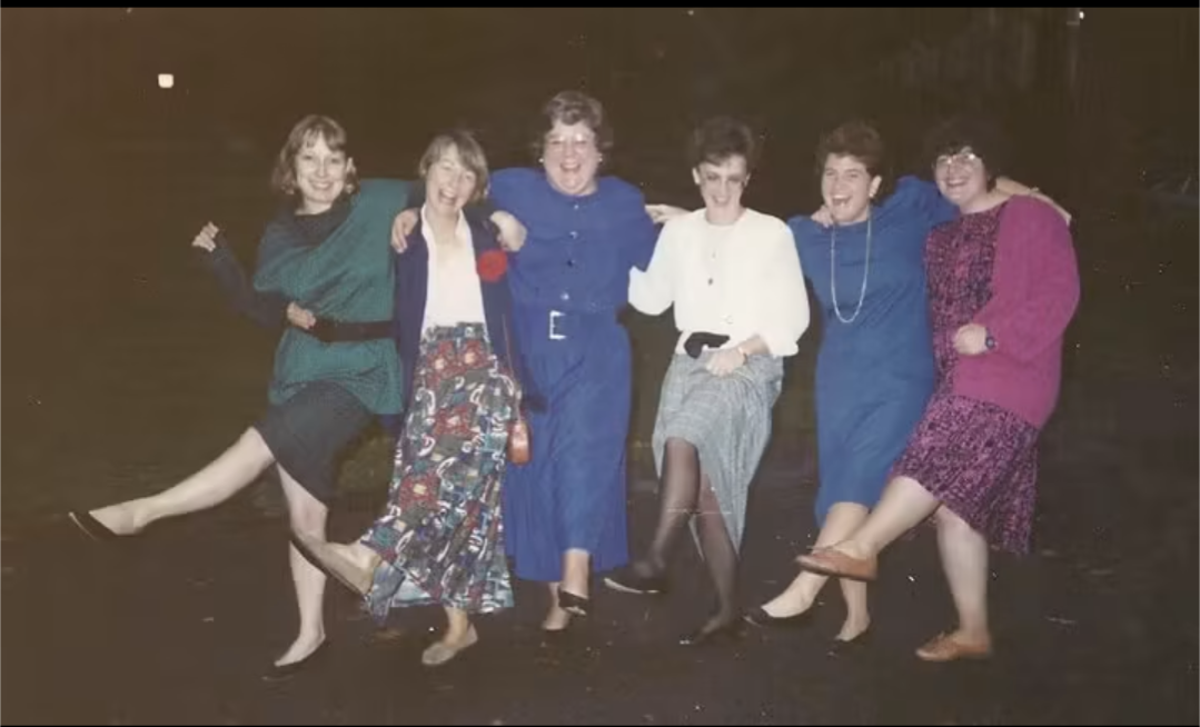 Six women standing outdoors at night, holding hands and smiling, with one leg raised. They wear colorful dresses and coats.