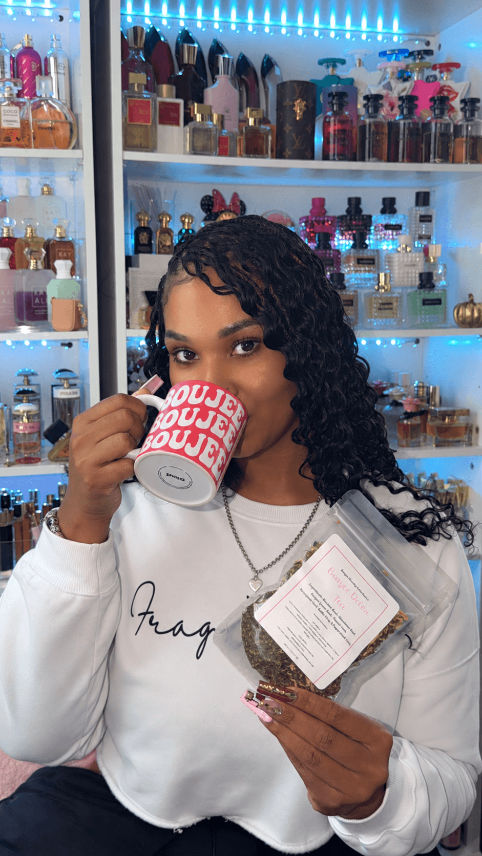 Young woman with curly hair drinking from a mug in a store with shelves of colorful bottles and products behind her.