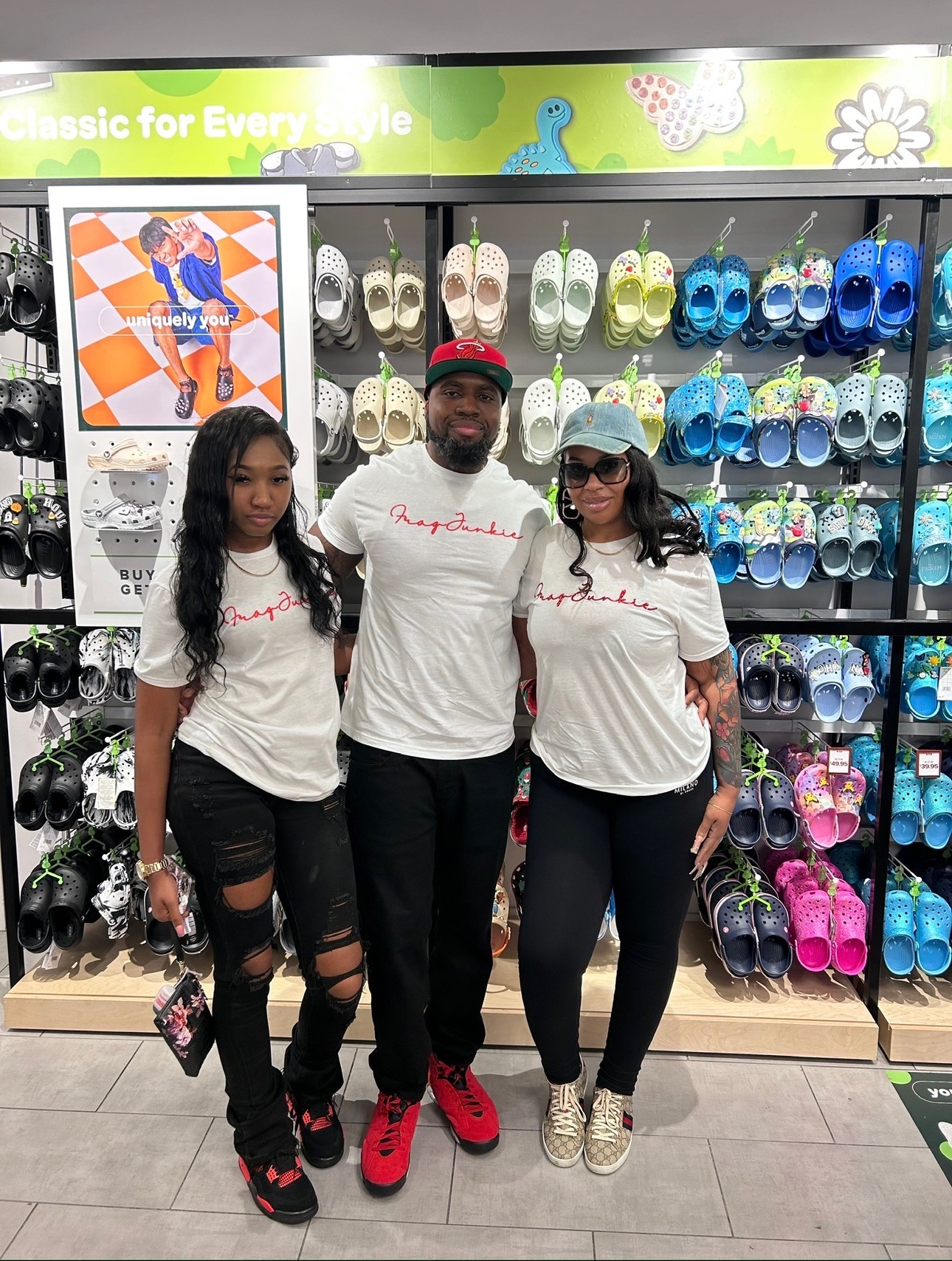 Three people stand in front of a shoe display, wearing matching white t-shirts, in a store with colorful footwear on shelves.