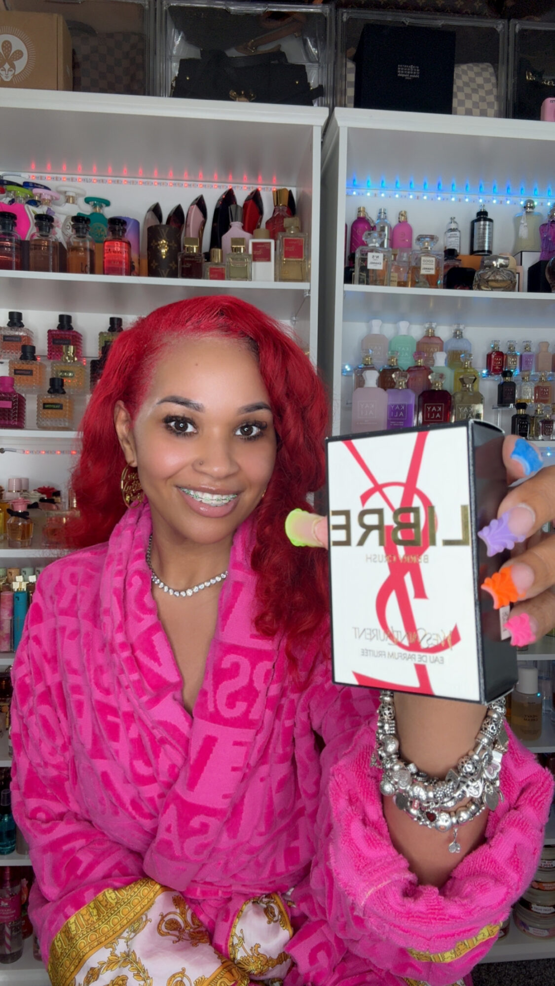 Young woman with red hair smiling, holding a phone with a box, in a nail salon with shelves of nail polish behind her.
