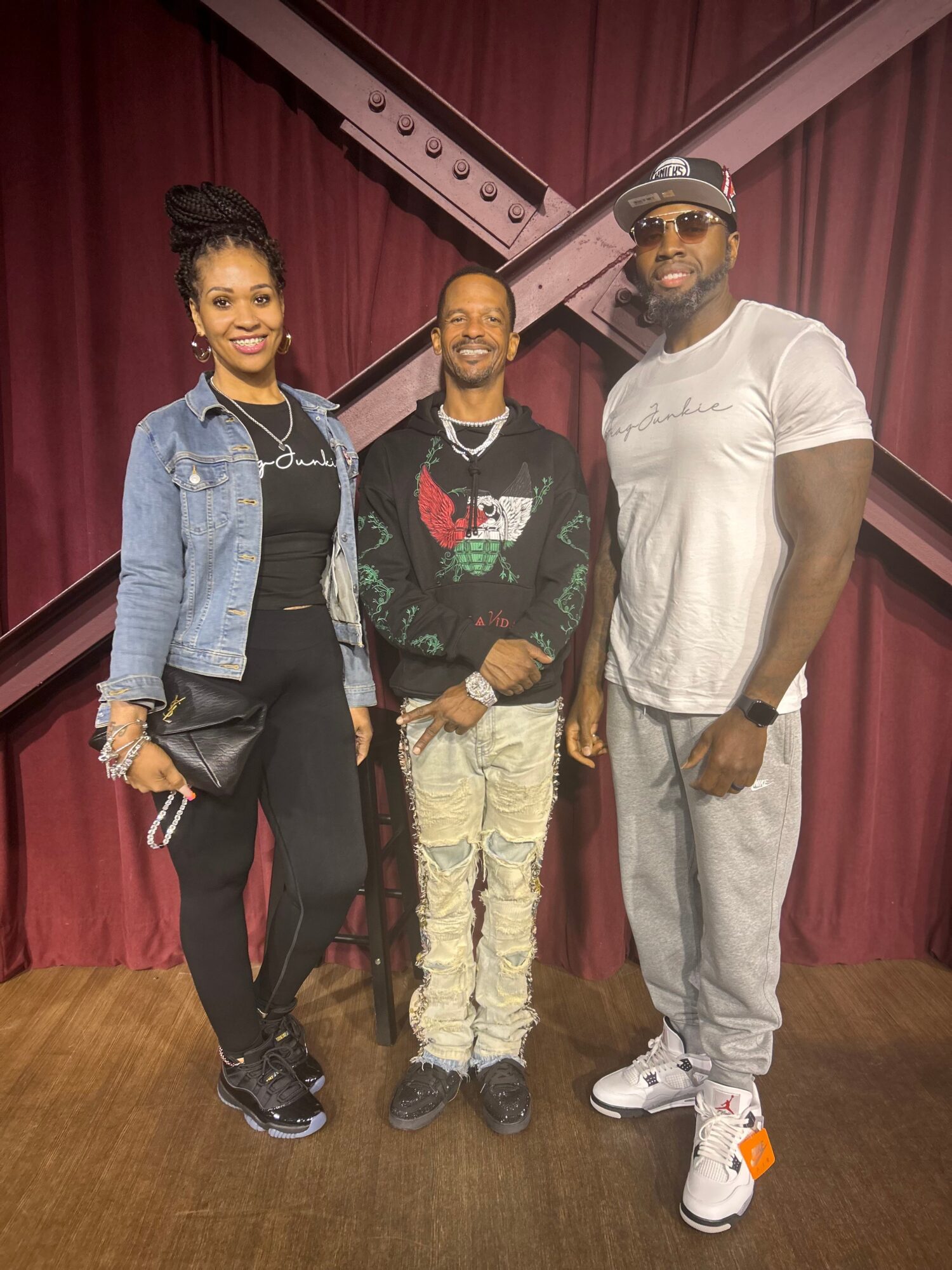 Three people standing in front of a maroon curtain, smiling, with casual clothing and accessories.